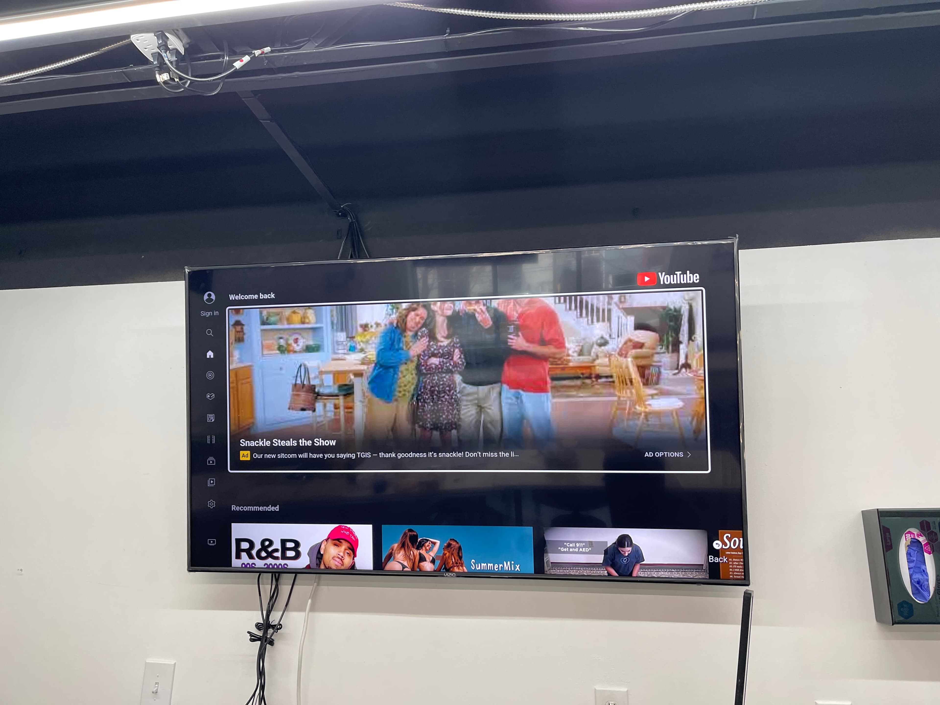 A television mounted on a wall displays a YouTube video, featuring people in a kitchen setting and a selection of recommended videos at the bottom.