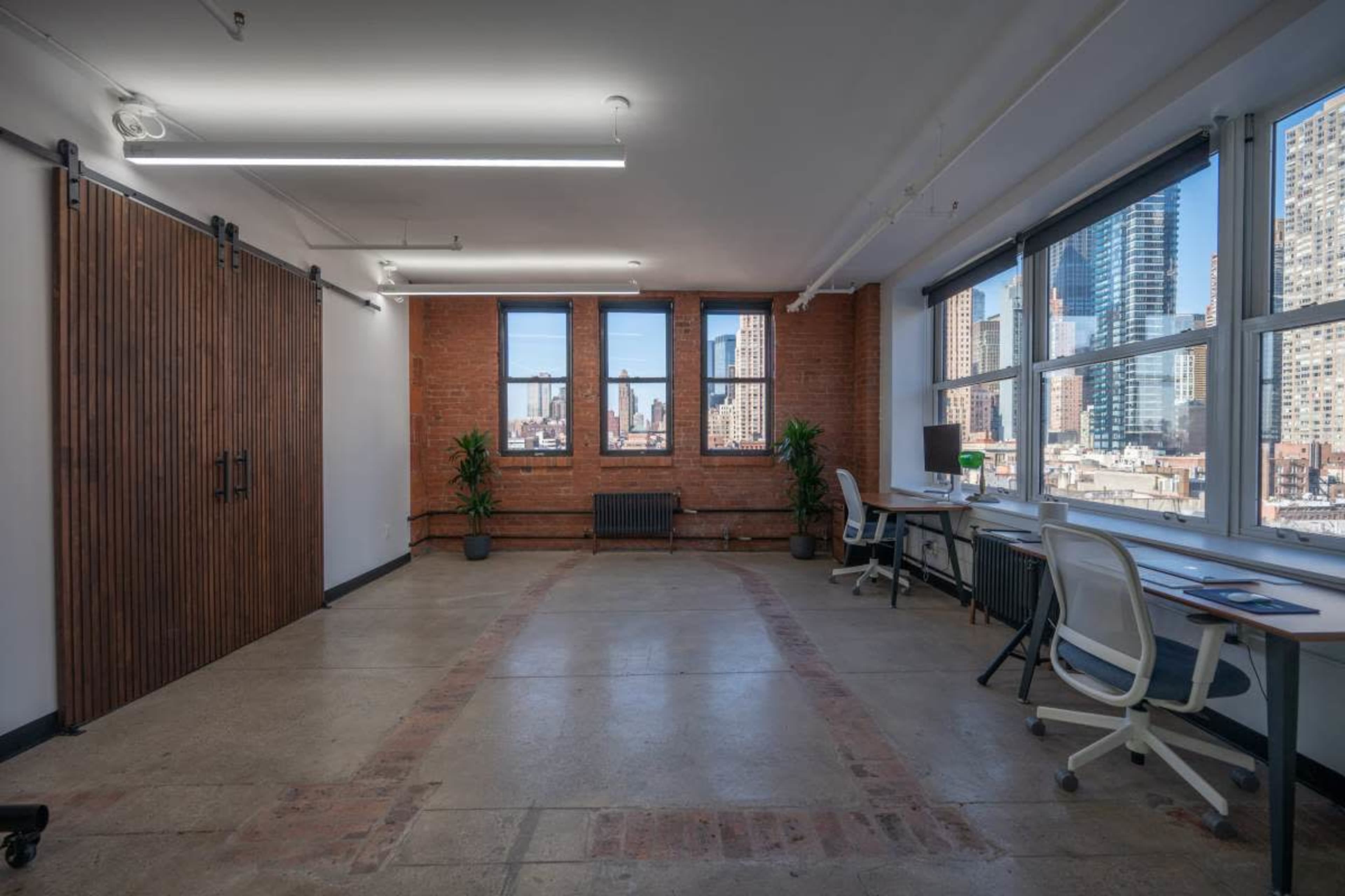The image shows a minimalist office space with large windows, exposed brick walls, and two workstations, featuring plants and wooden sliding doors.