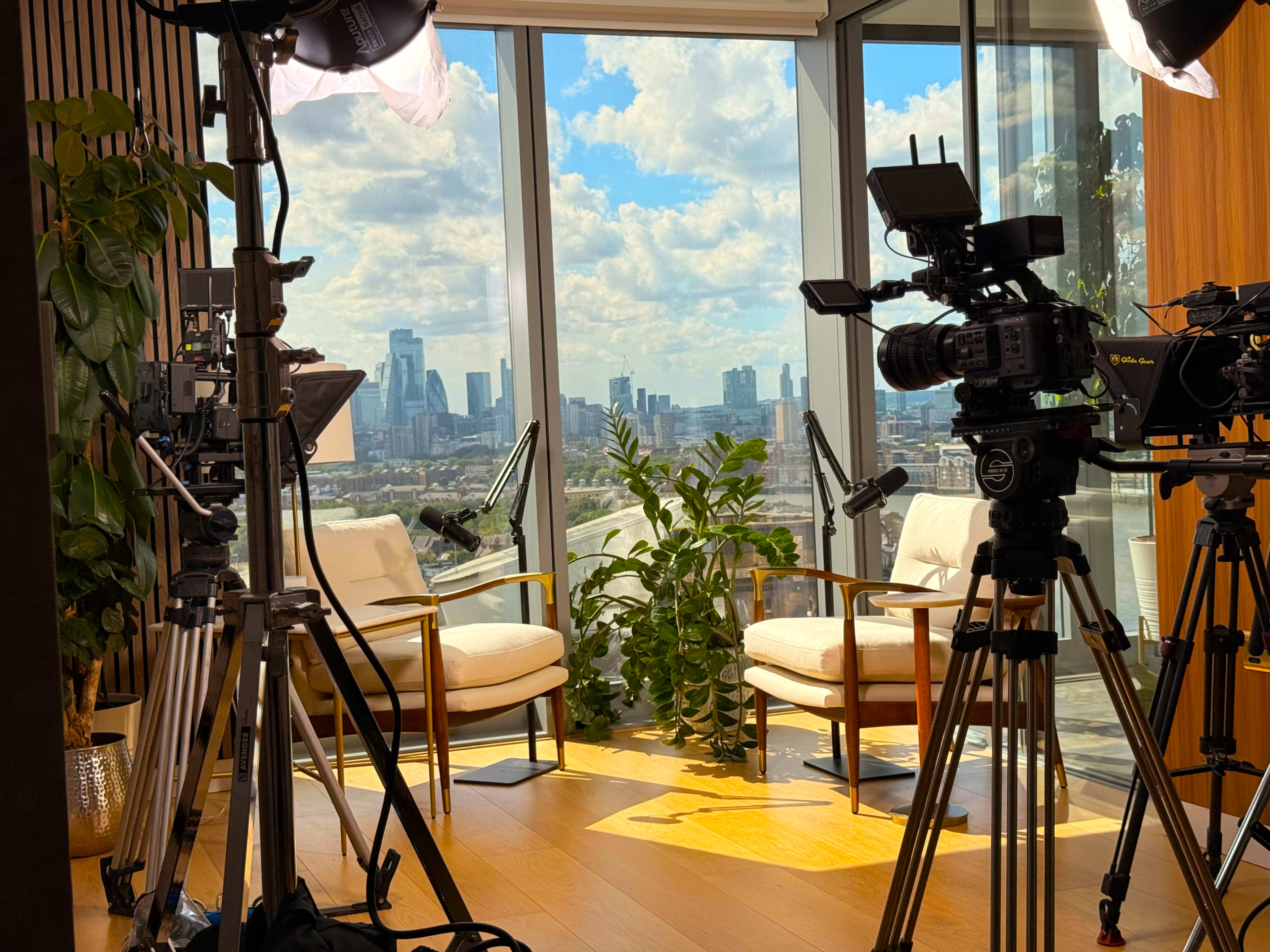 The image shows a well-lit room with two chairs and a view of a city skyline through large windows, flanked by video cameras and lighting equipment.