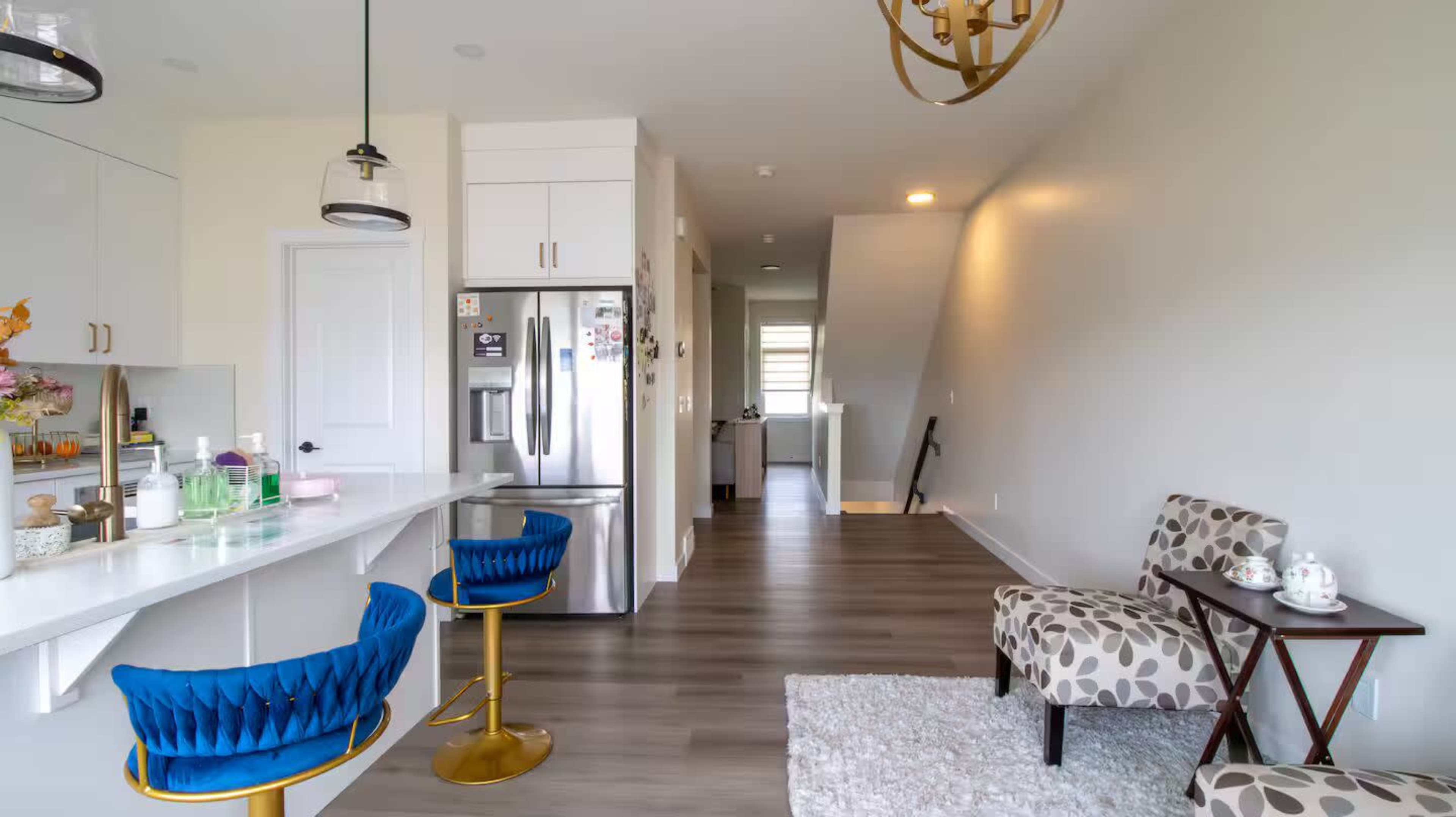 The image shows a modern kitchen and living area with blue chairs at a breakfast bar, a stainless steel refrigerator, and a staircase leading to another level.