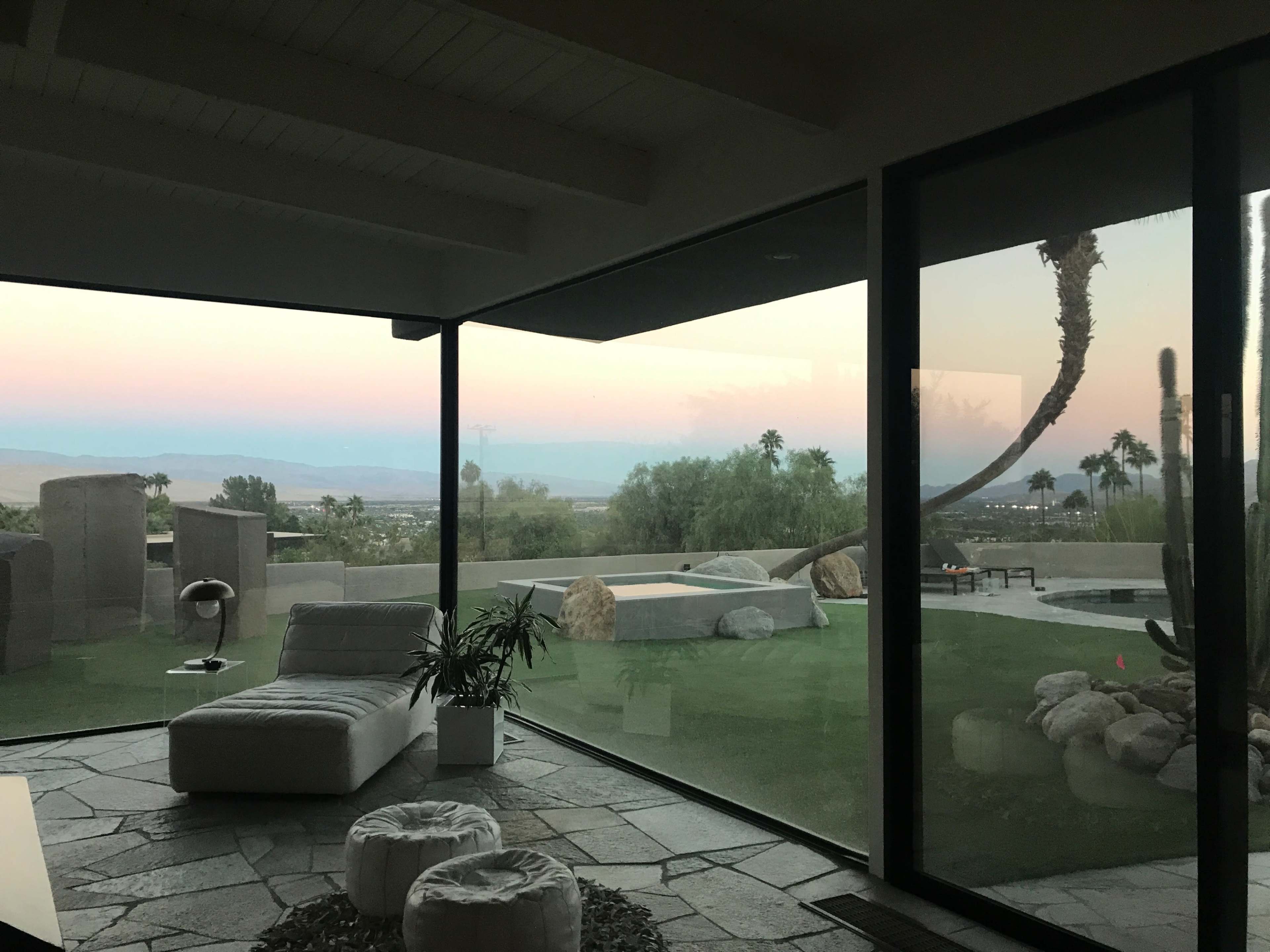 A modern living space with large glass walls overlooking a landscaped outdoor area featuring palm trees and a soft sunset sky.