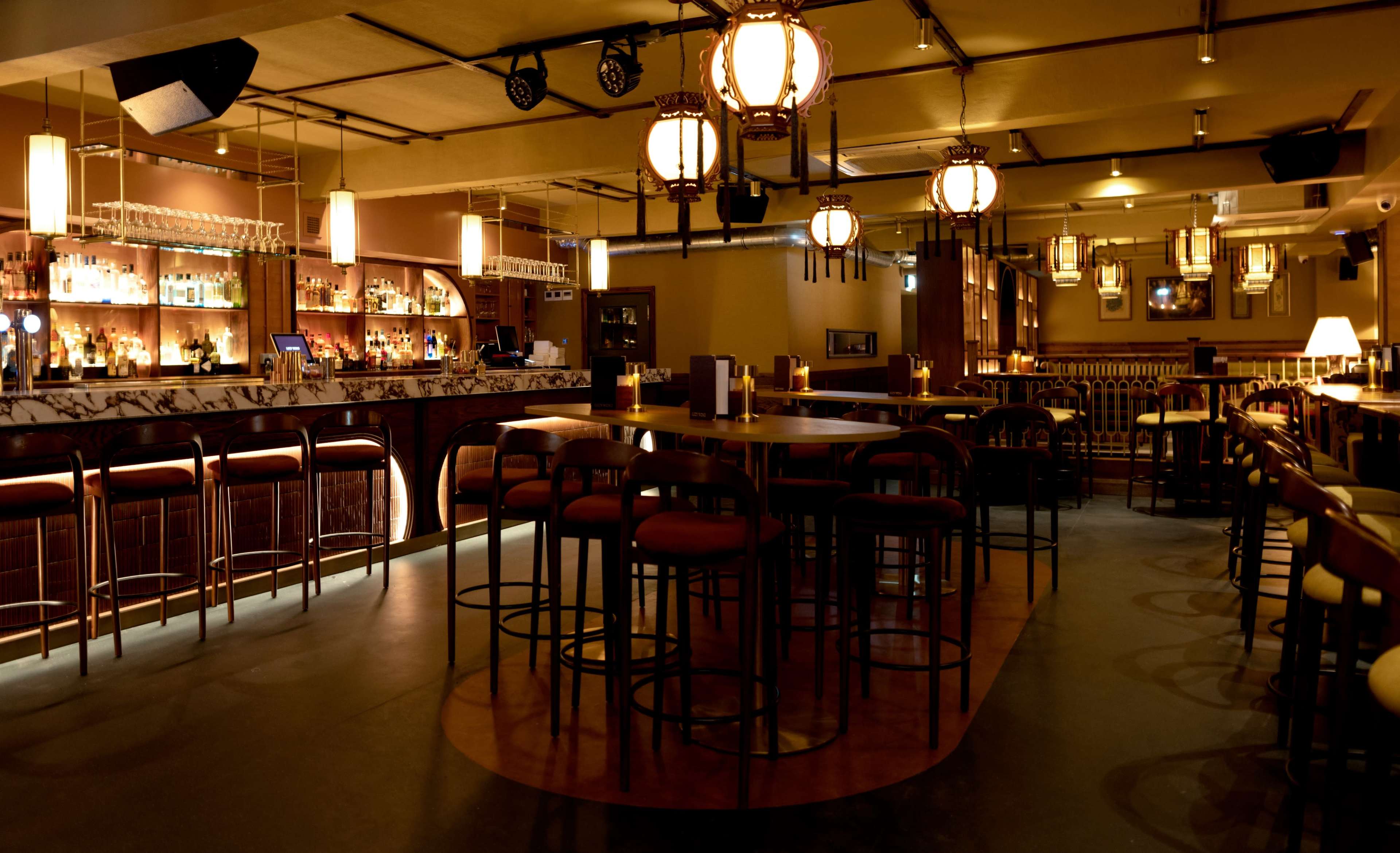 A dimly lit bar features wooden furnishings, pendant lights, and a polished countertop with an array of liquor bottles on display.