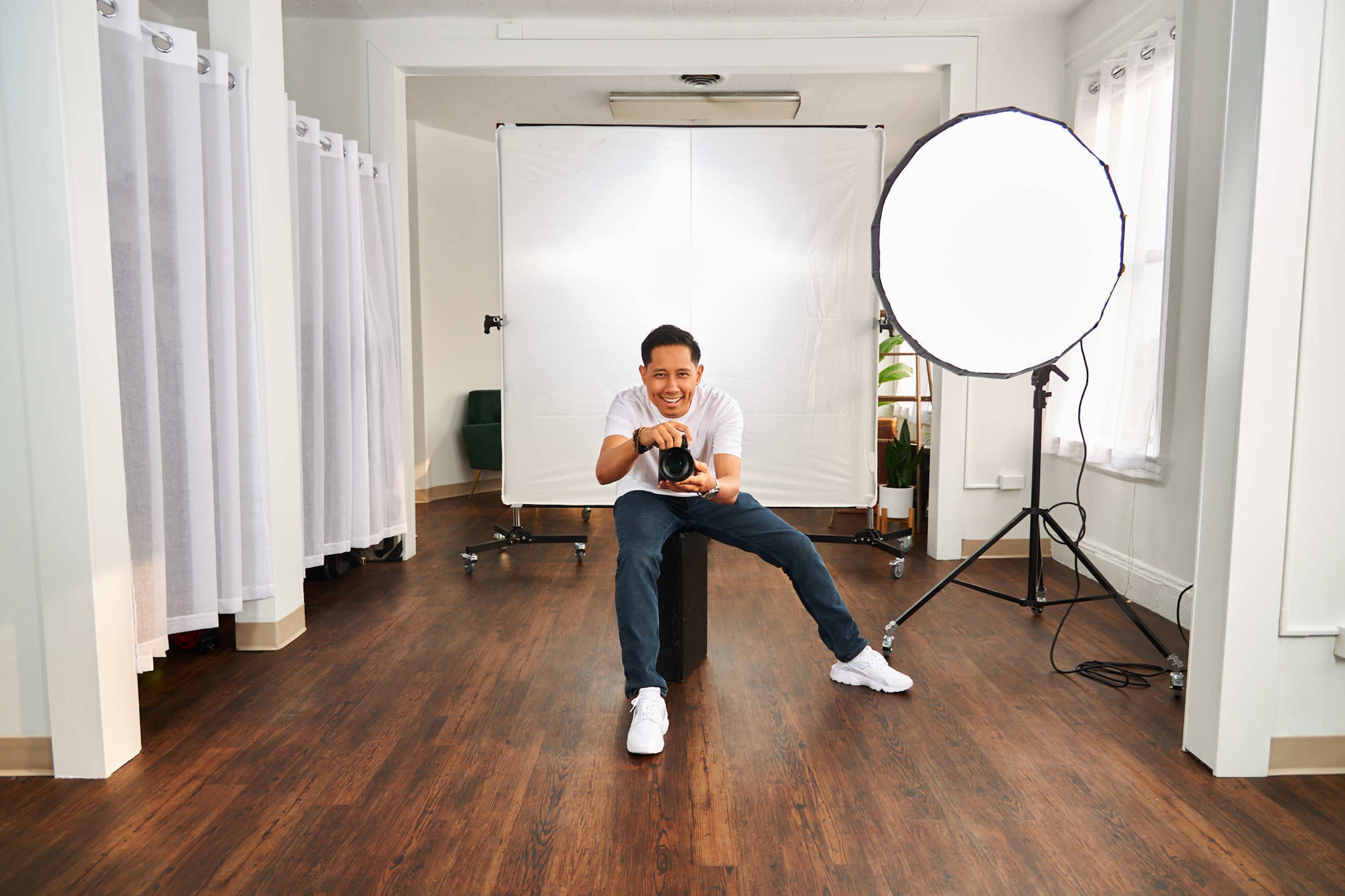A person sits on a block in a well-lit studio, holding a camera and smiling, with photo backdrop equipment and a softbox nearby.