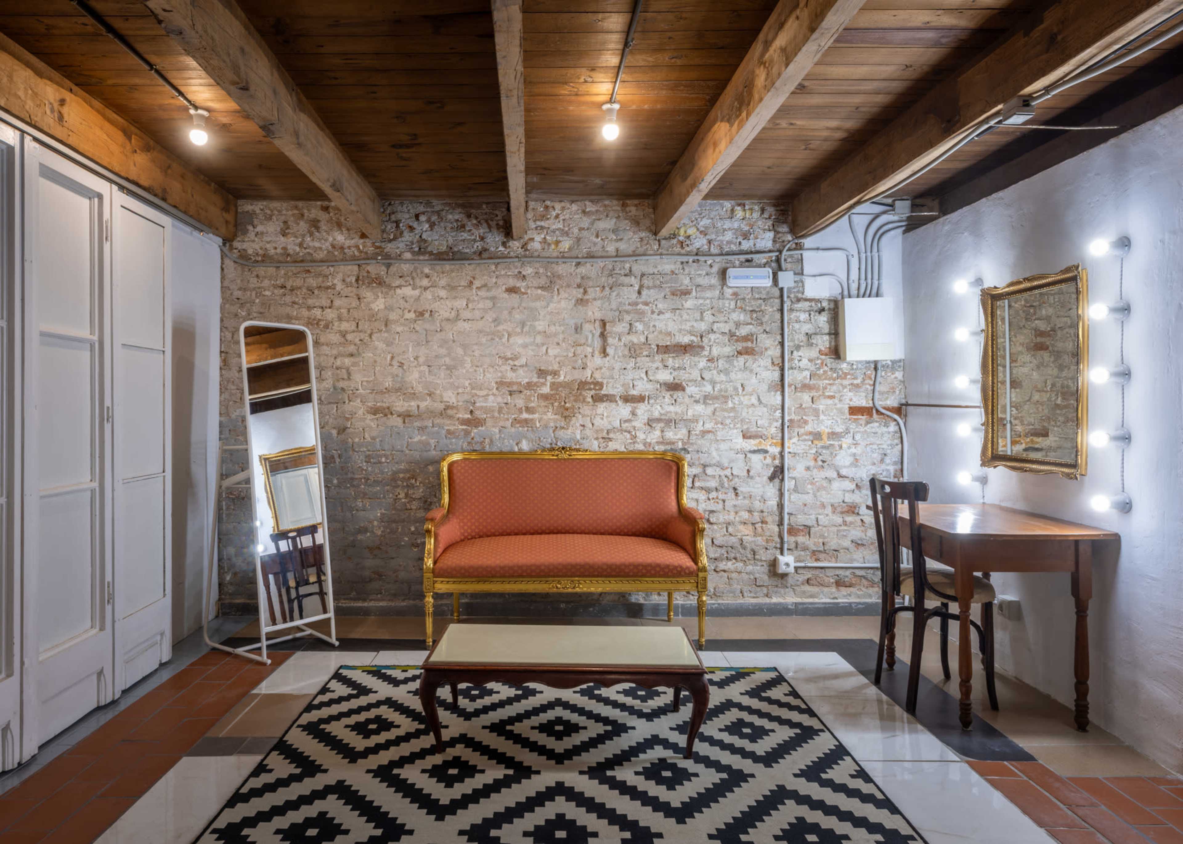 The image shows a well-lit, minimalist room with exposed brick walls, featuring a vintage orange sofa, a wooden table, and a large mirror beside a dressing table.