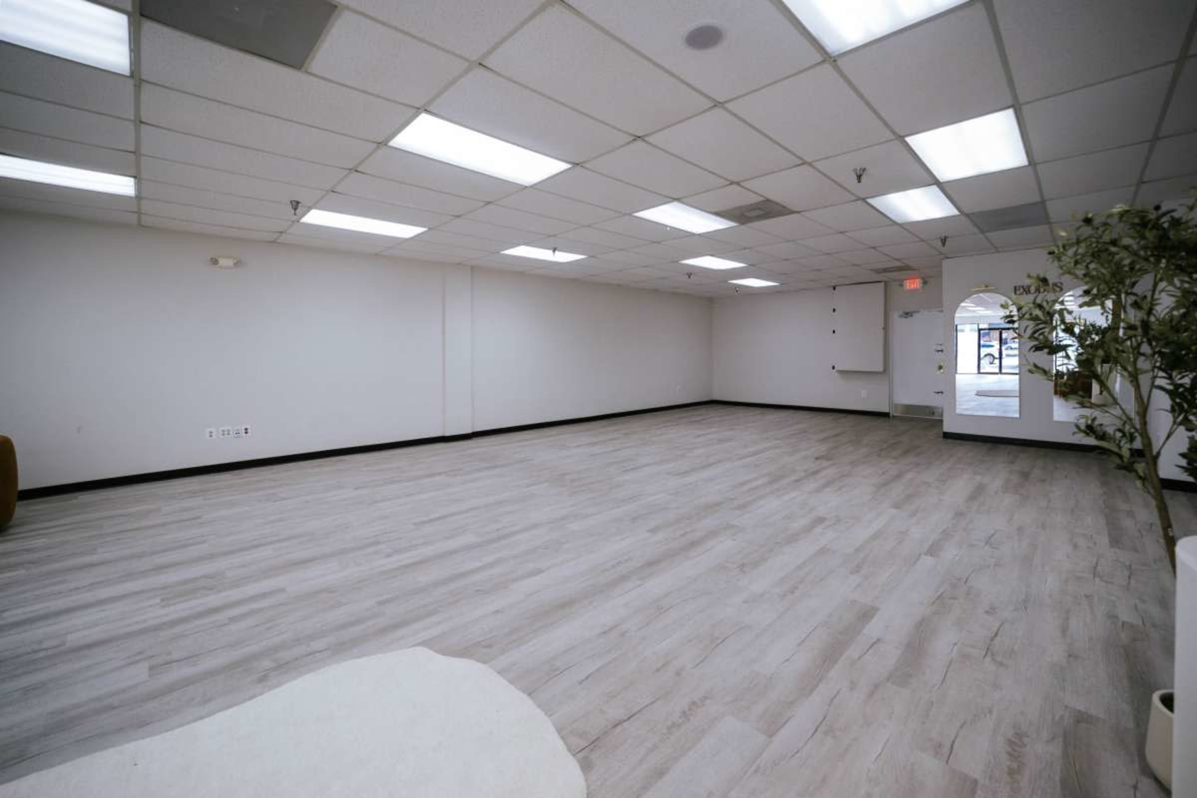 The image shows an empty, well-lit room with a wooden floor and a single door leading outside.