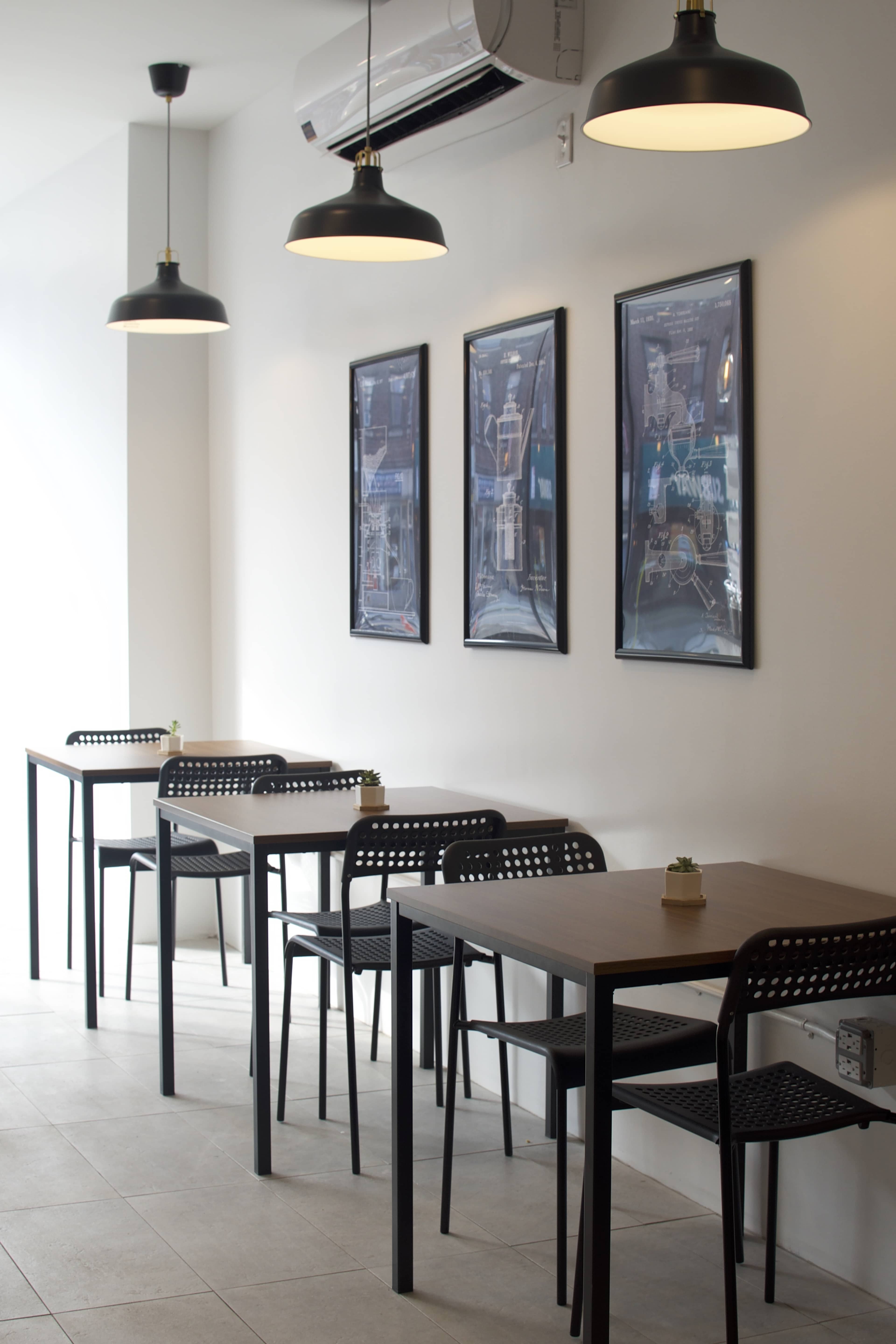 A minimalist café interior with several black tables and chairs arranged along a brightly lit wall adorned with framed pictures.