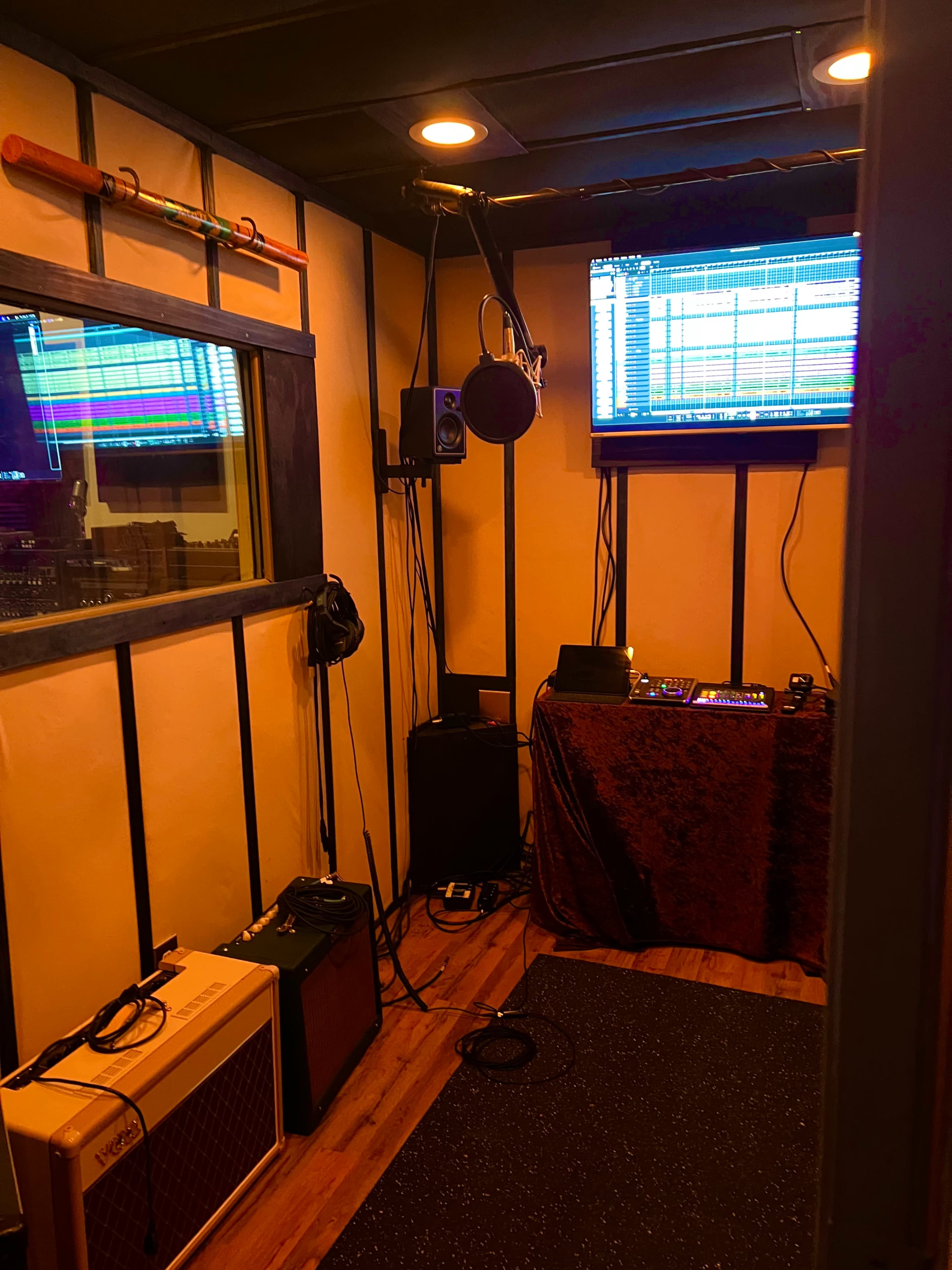 The image shows a small recording studio with soundproof walls, a microphone setup, a display screen showing audio tracks, and various musical equipment.