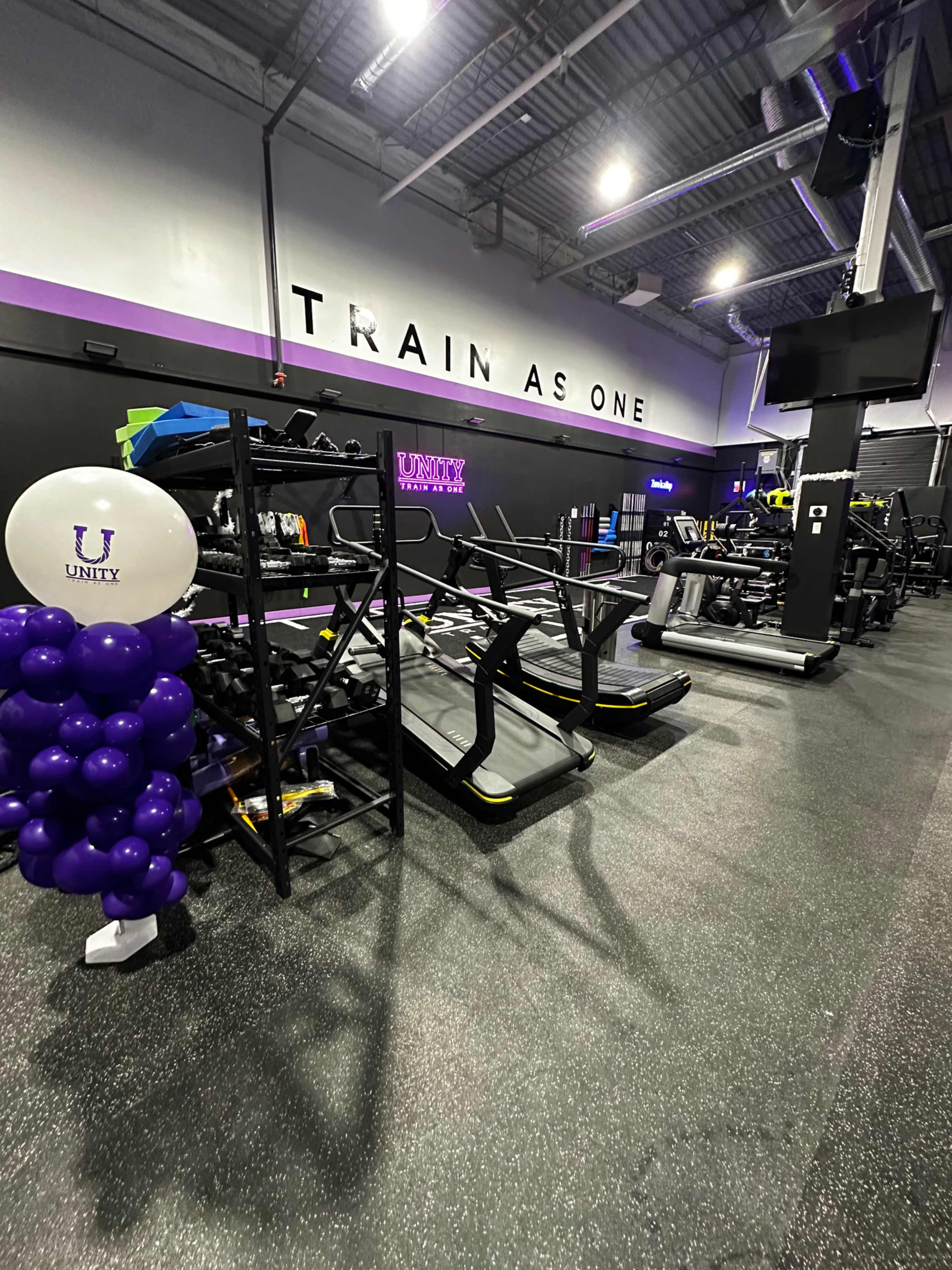 A gym interior with exercise equipment lined up along the wall, a balloon display, and the phrase "TRAIN AS ONE" prominently on the wall.