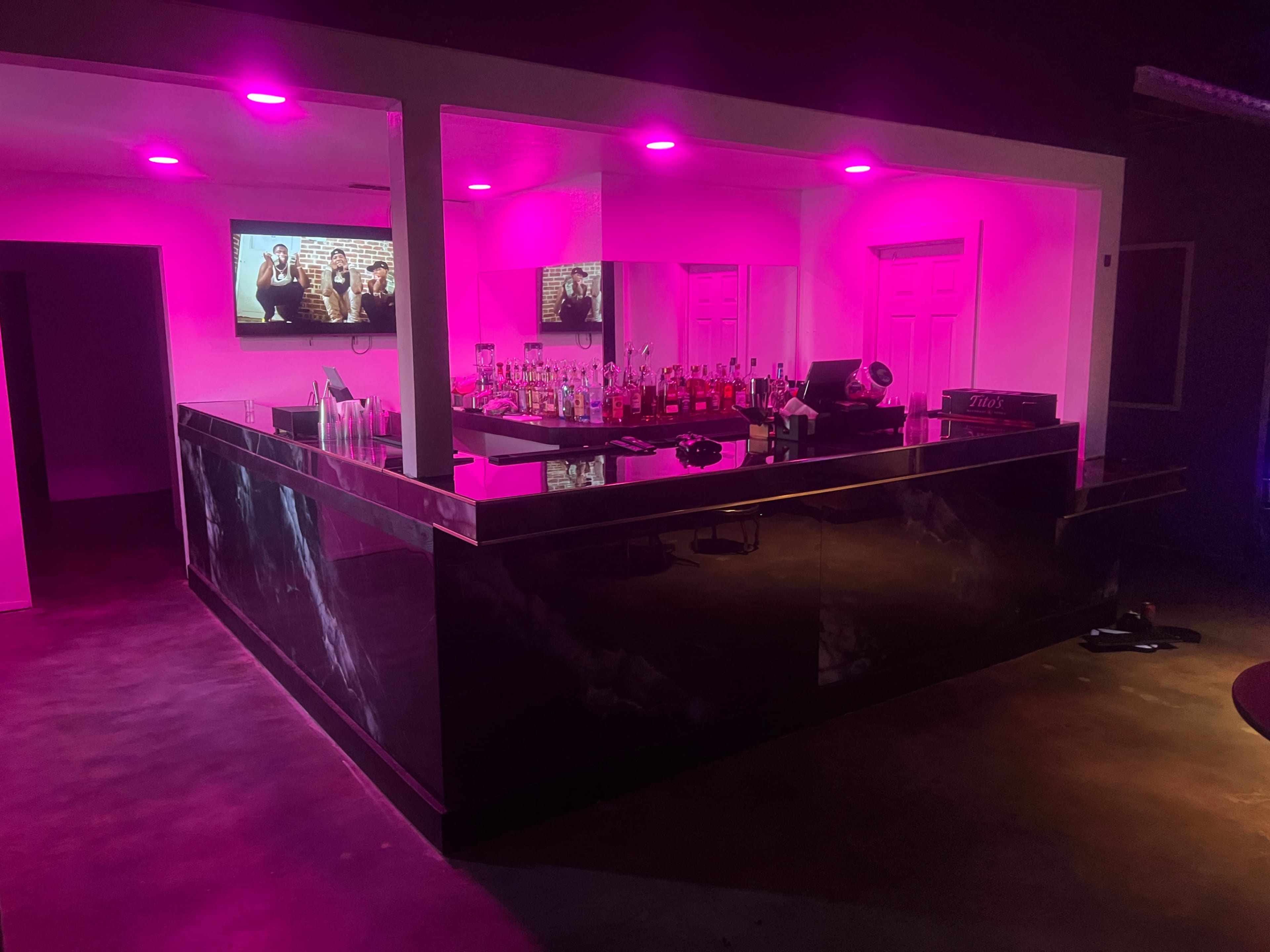 The image shows a bar area illuminated with pink lighting, featuring a counter with various bottles behind it and a television mounted on the wall.