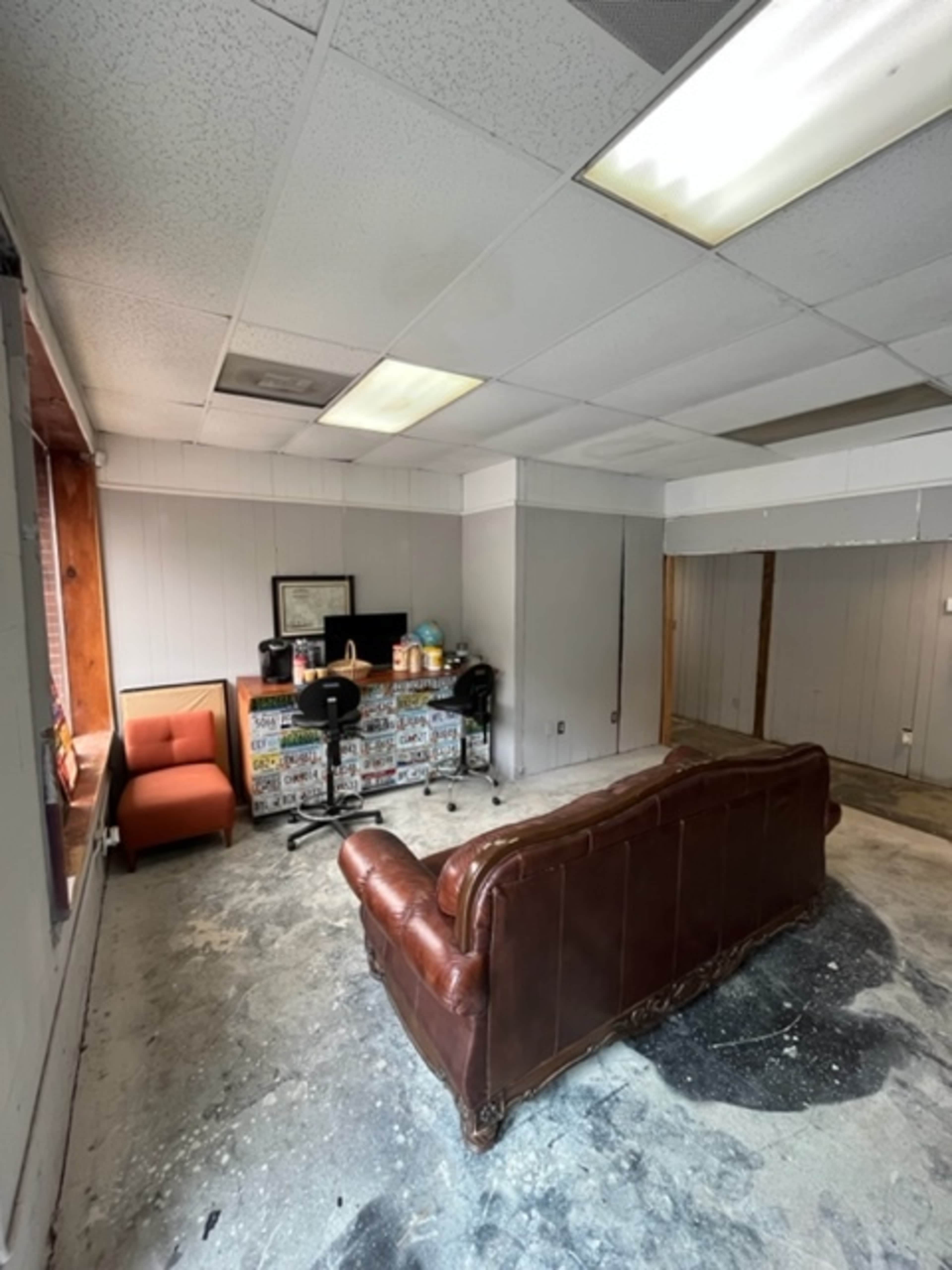 The image shows a room with a brown leather couch, an orange chair, two black chairs at a desk covered with stickers, and gray walls, all under a ceiling with fluorescent lights.