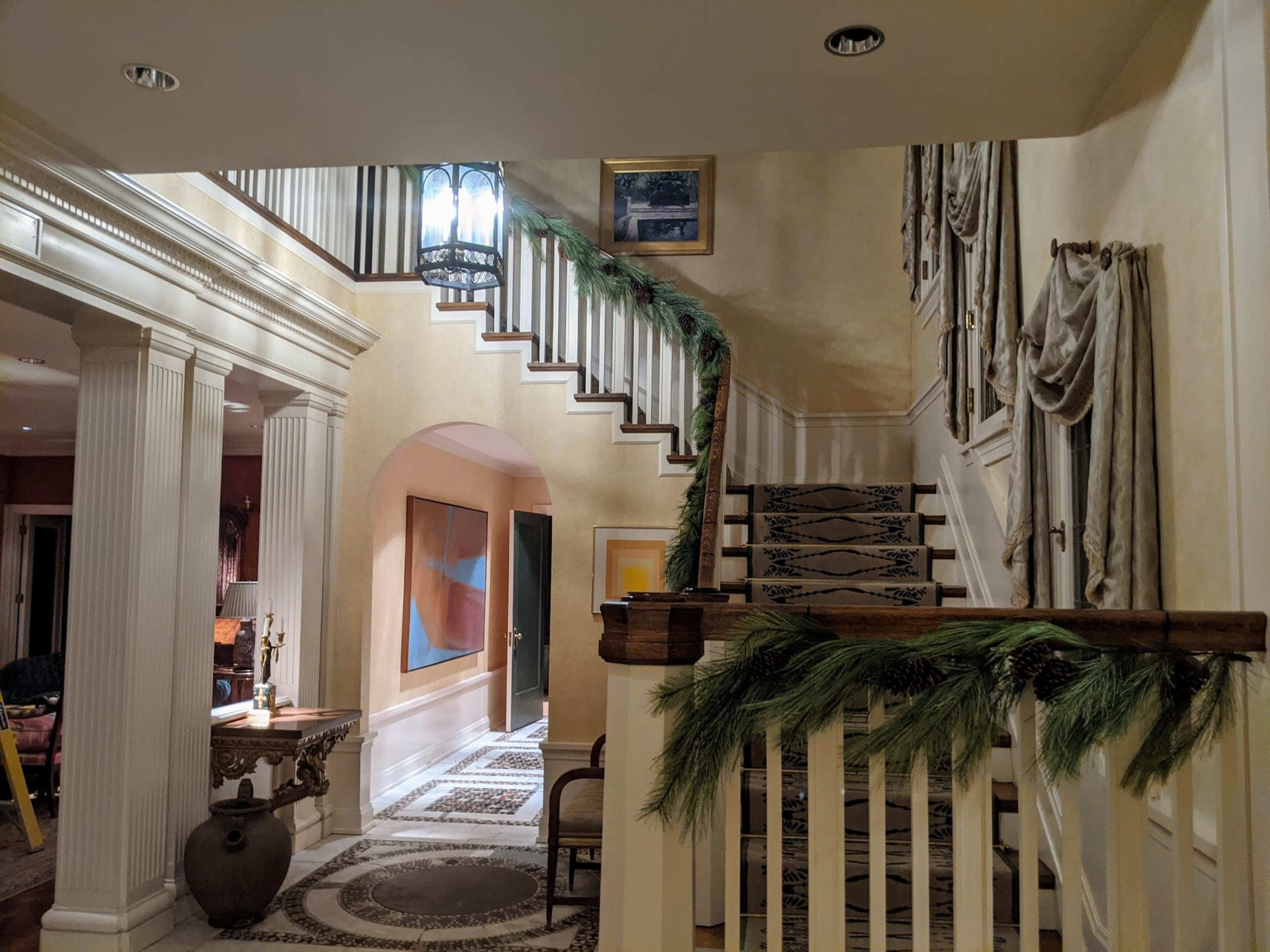 The image shows a well-decorated foyer with a staircase adorned with greenery, featuring a chandelier and artwork on the walls.
