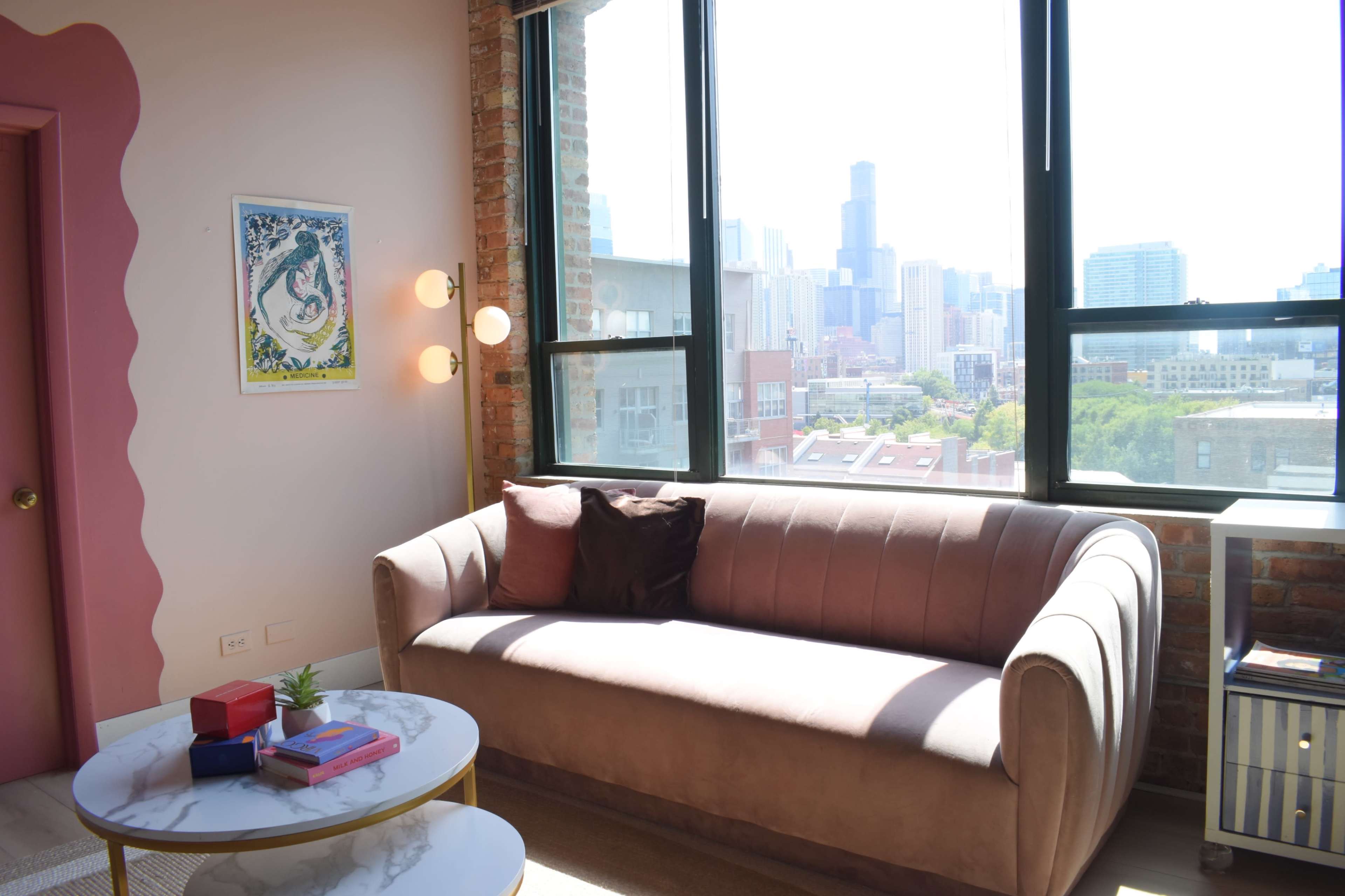 A modern living room features a pink sofa, a round marble coffee table with books and a small plant, and large windows overlooking a city skyline.