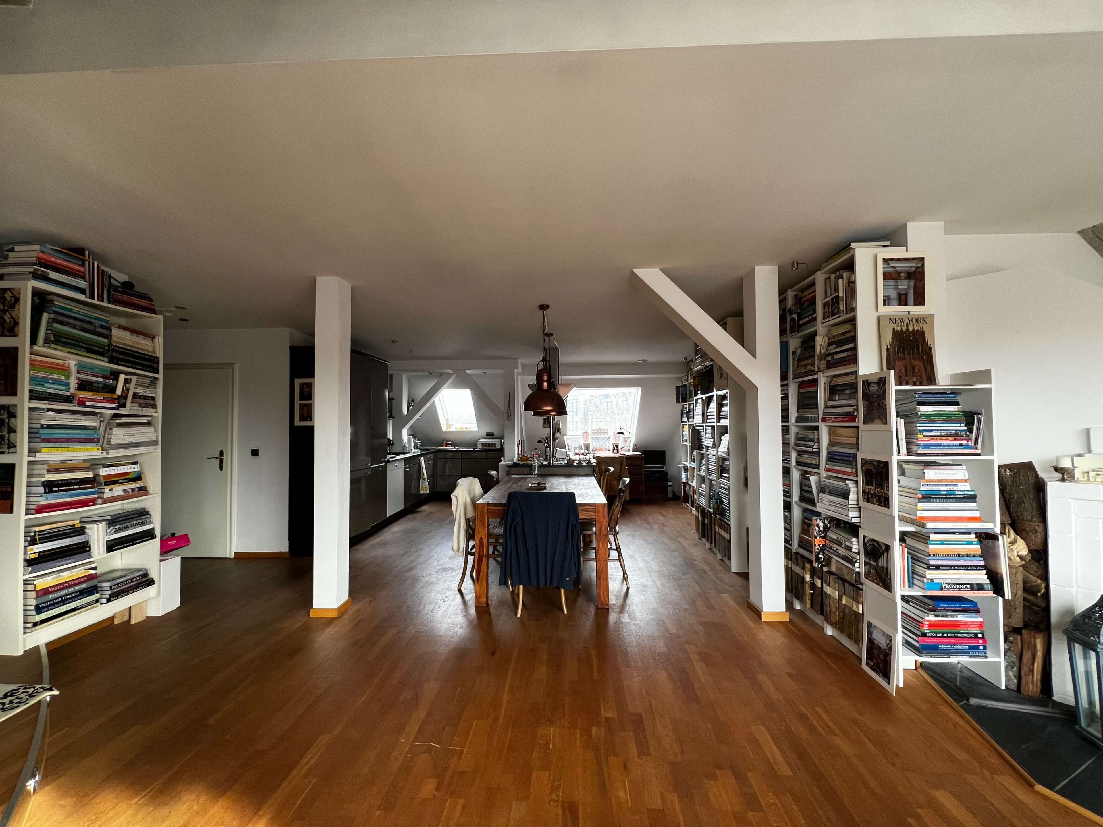 The image shows a spacious living area with wooden flooring, a long dining table at the center, and bookshelves filled with books lining the walls.
