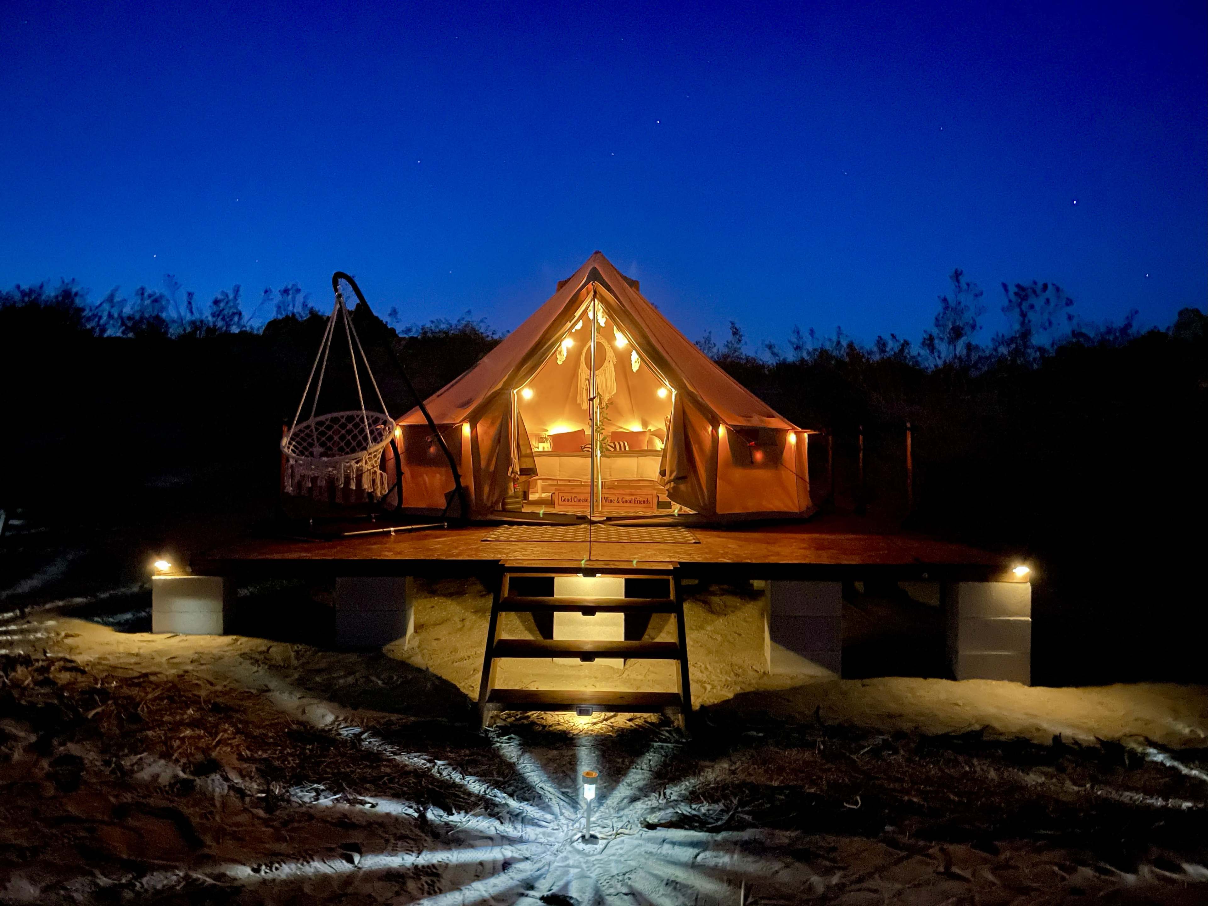 A glamping tent with warm lights stands on a wooden deck at night, surrounded by subtle landscaping and an ambient glow from ground lights.