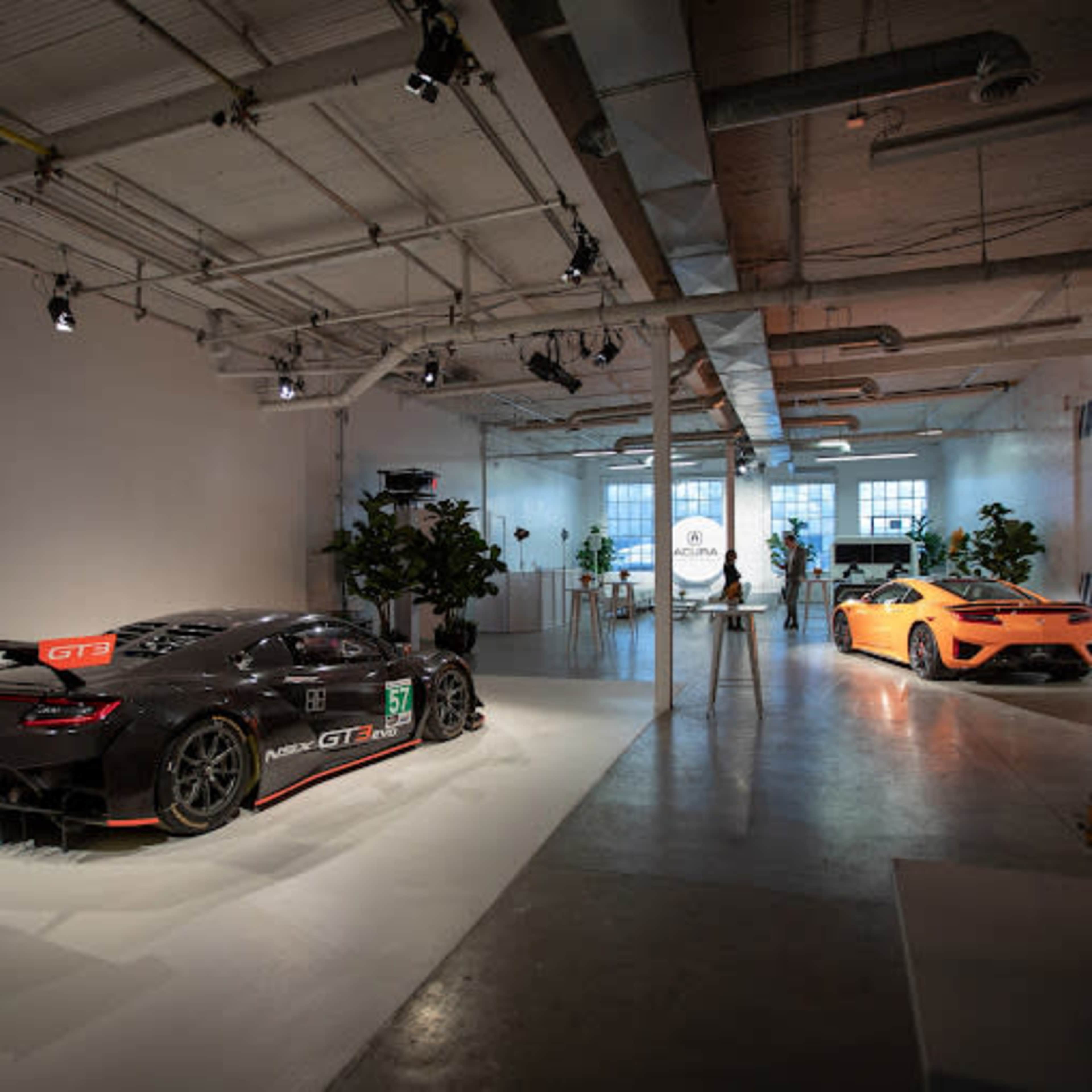 Two high-performance sports cars, one black and one orange, positioned in a well-lit, minimalist showroom with potted plants and a sign in the background.