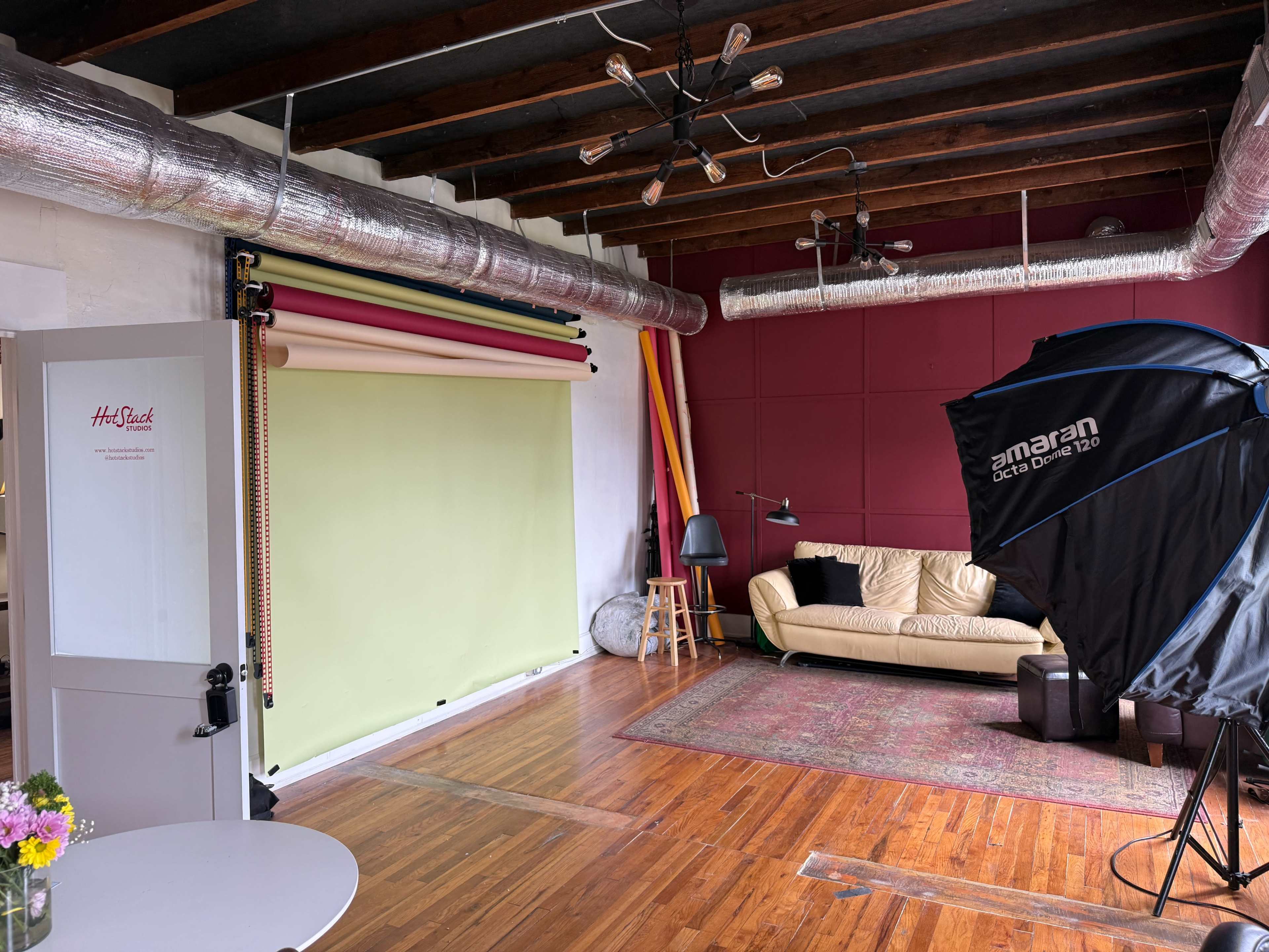 The image depicts a photography studio with a green backdrop, wooden flooring, a sofa, and various studio equipment.