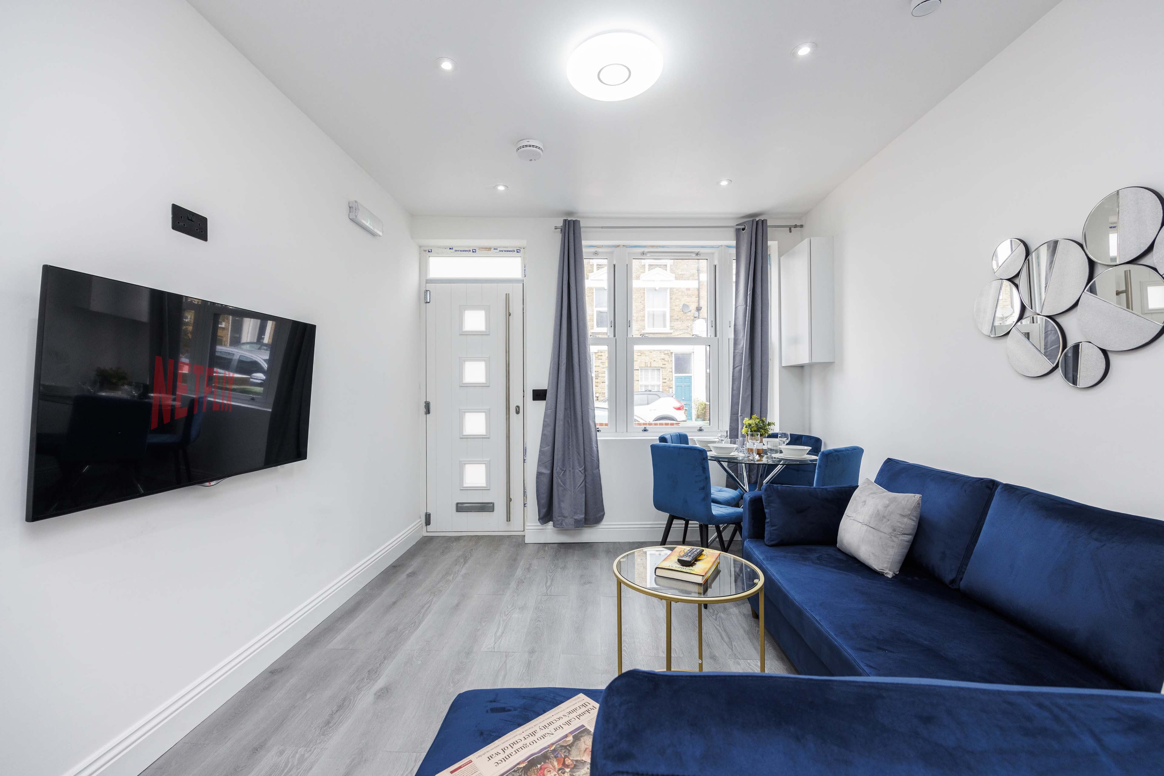 A modern living room features a blue sofa, a round coffee table, a flat-screen TV mounted on the wall, and a window with curtains.