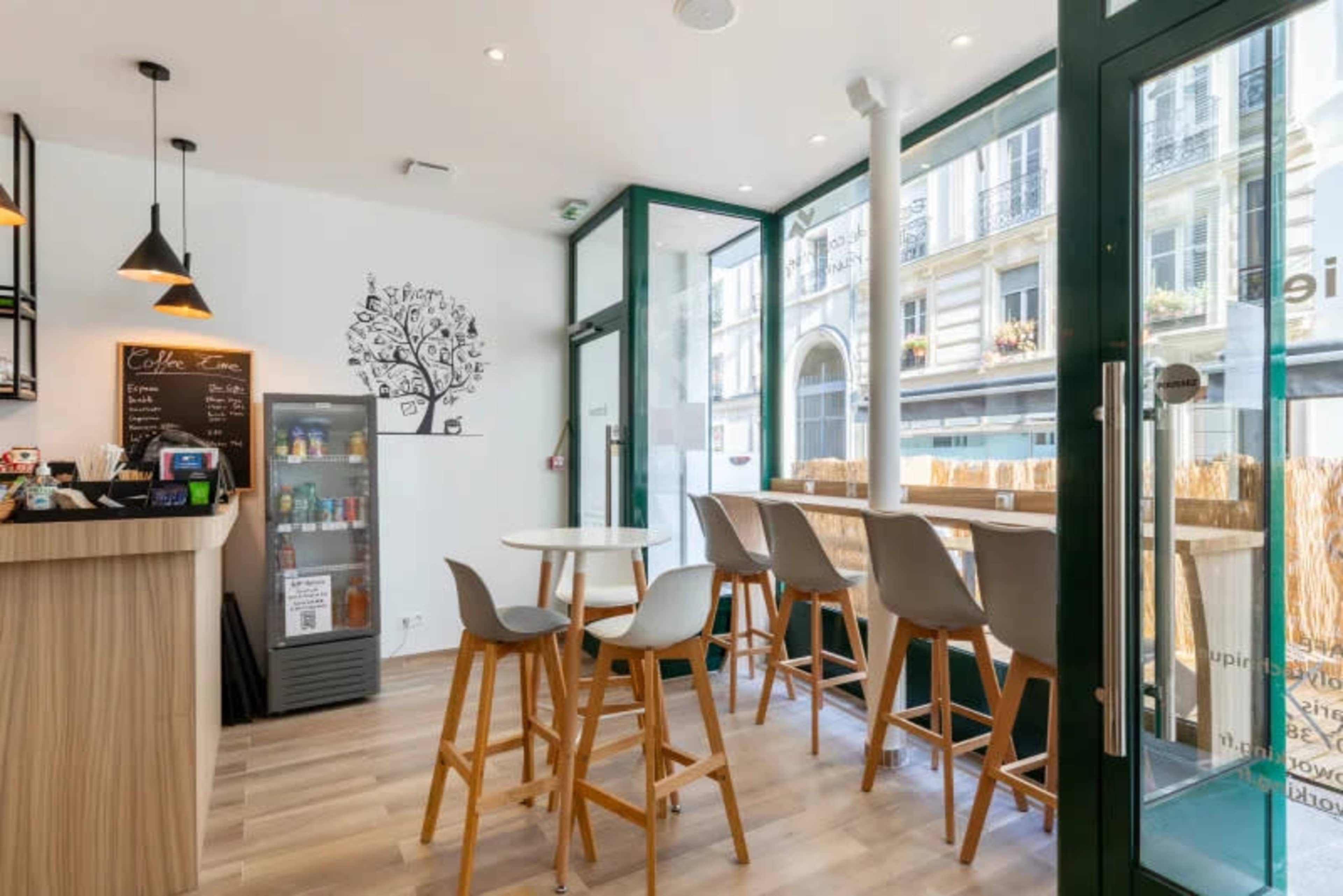 The image shows a modern café interior with a minimalist design, featuring a counter, a refrigerator, and high chairs at windows overlooking the street.