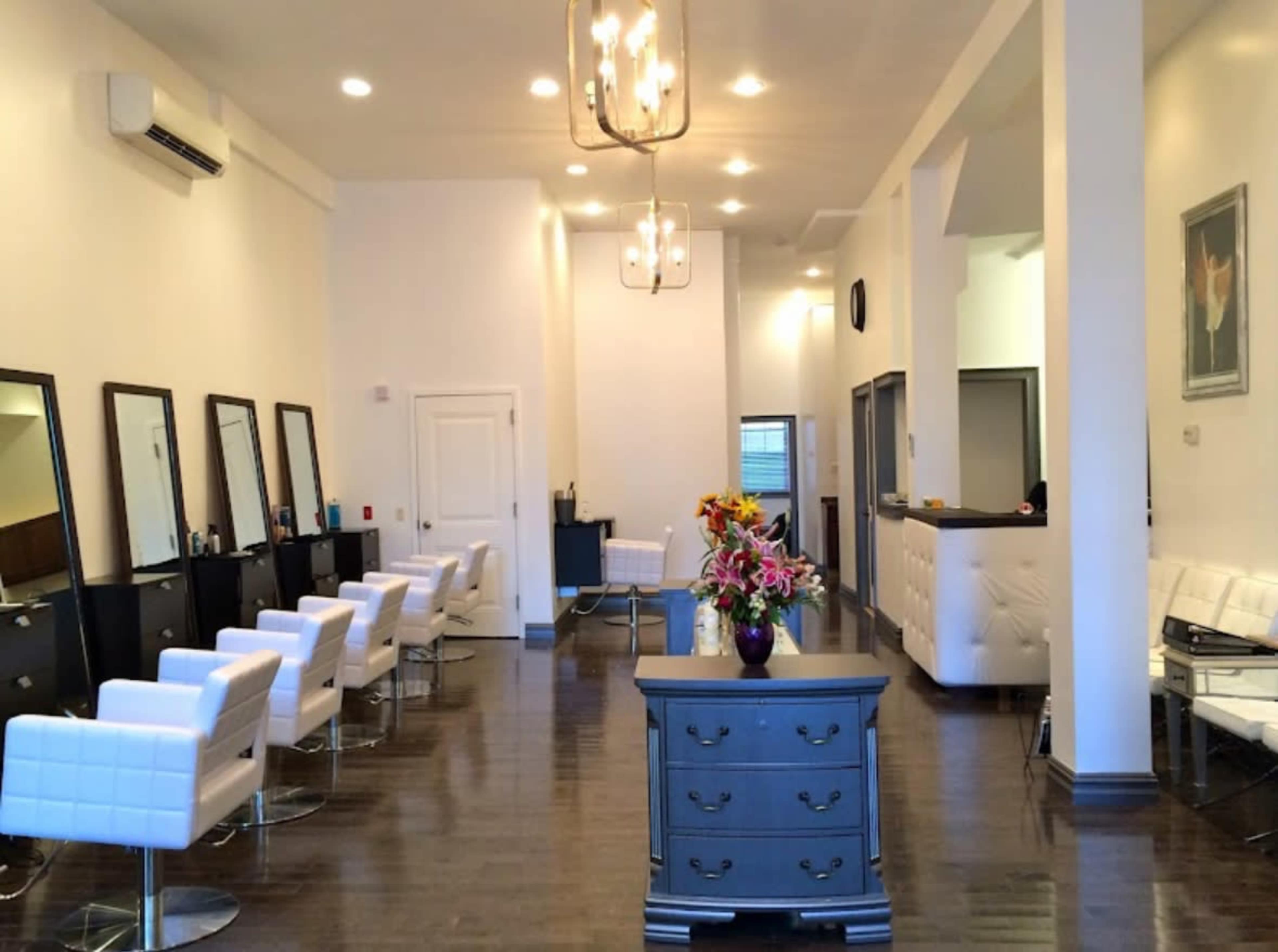 A modern hair salon features several styling stations with white chairs and large mirrors, along with a central reception area and floral arrangements on a gray chest of drawers.