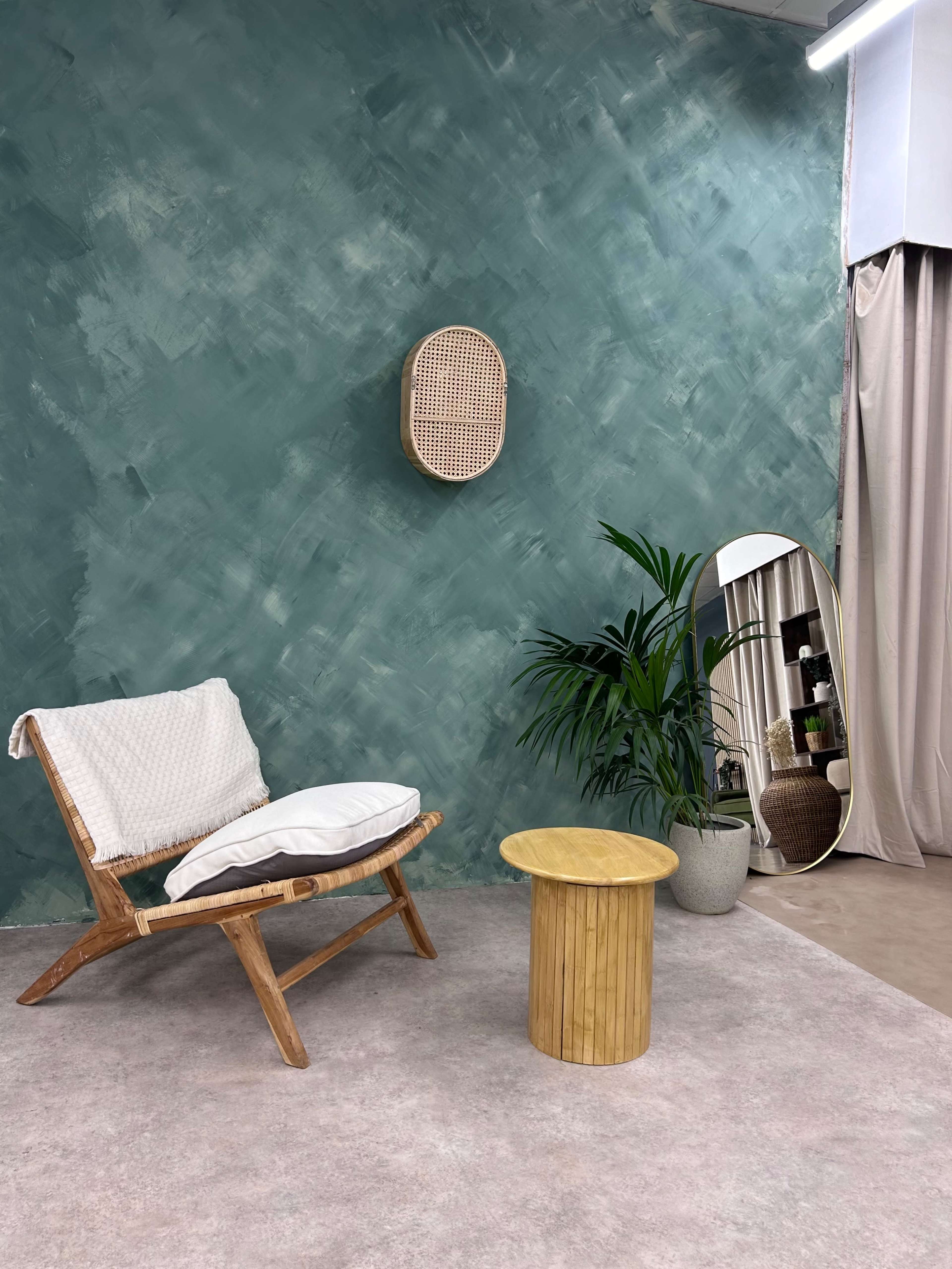 A minimalist interior space featuring a wooden armchair with a cushion, a small yellow table, a potted plant, and a tall mirror against a textured teal wall.