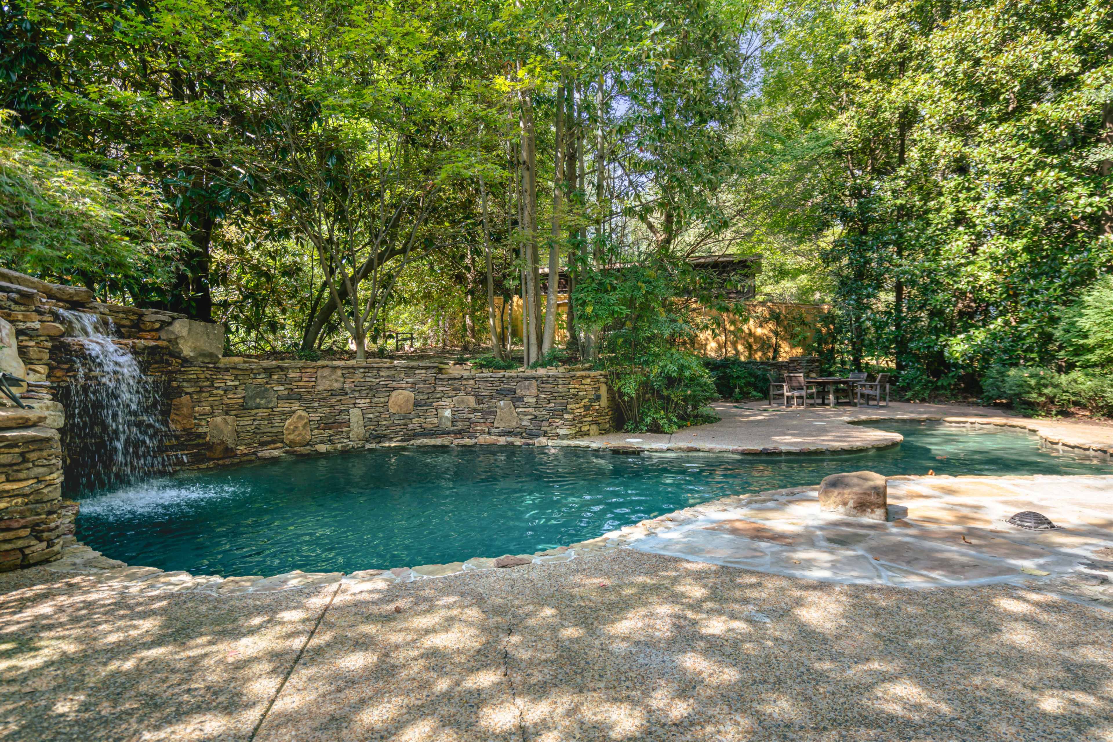 The image shows a naturalistic swimming pool surrounded by stone features, a small waterfall, and lush greenery.