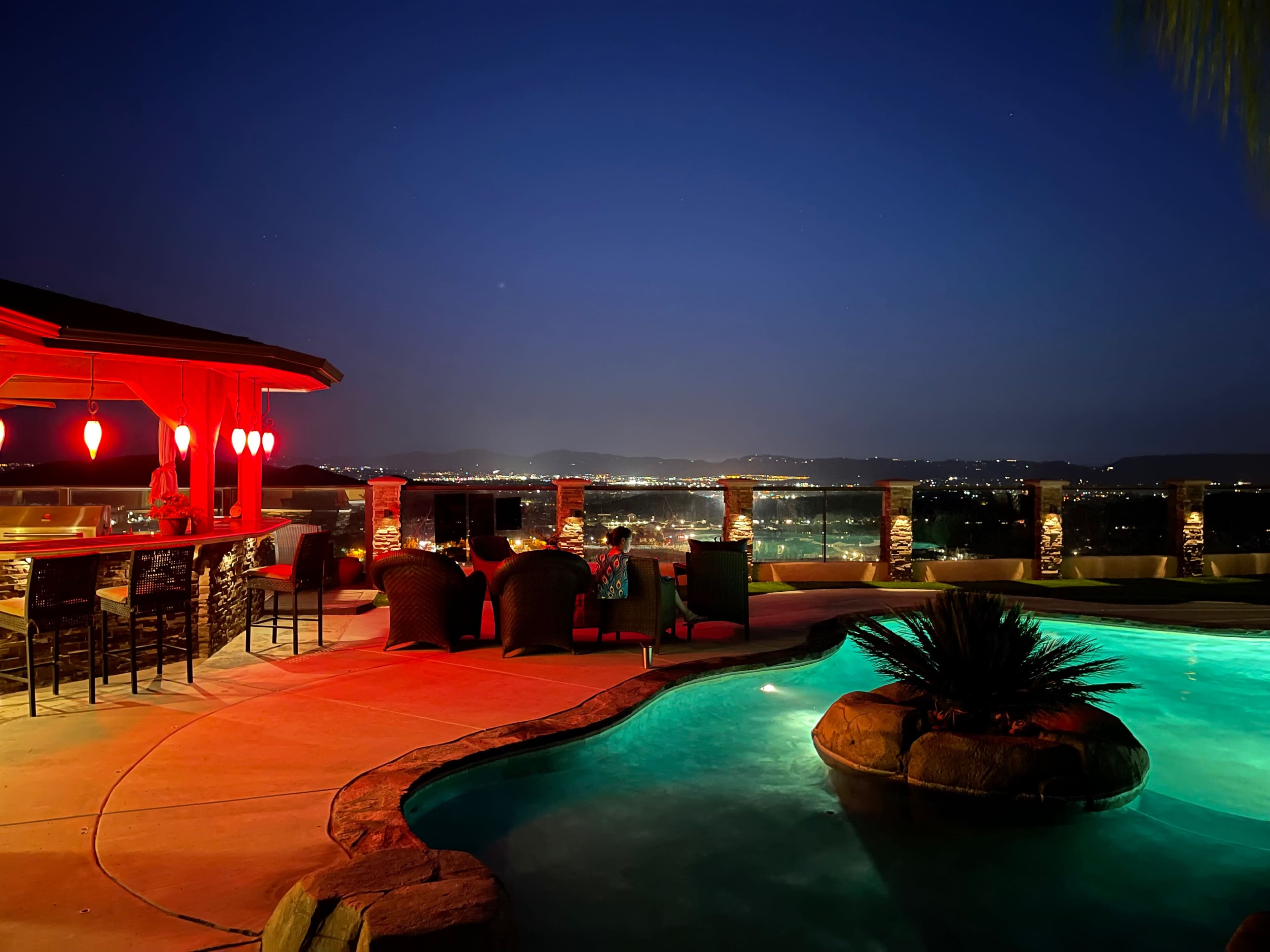 A luxurious backyard patio features a lit pool, a bar area, and panoramic views of the city at night.