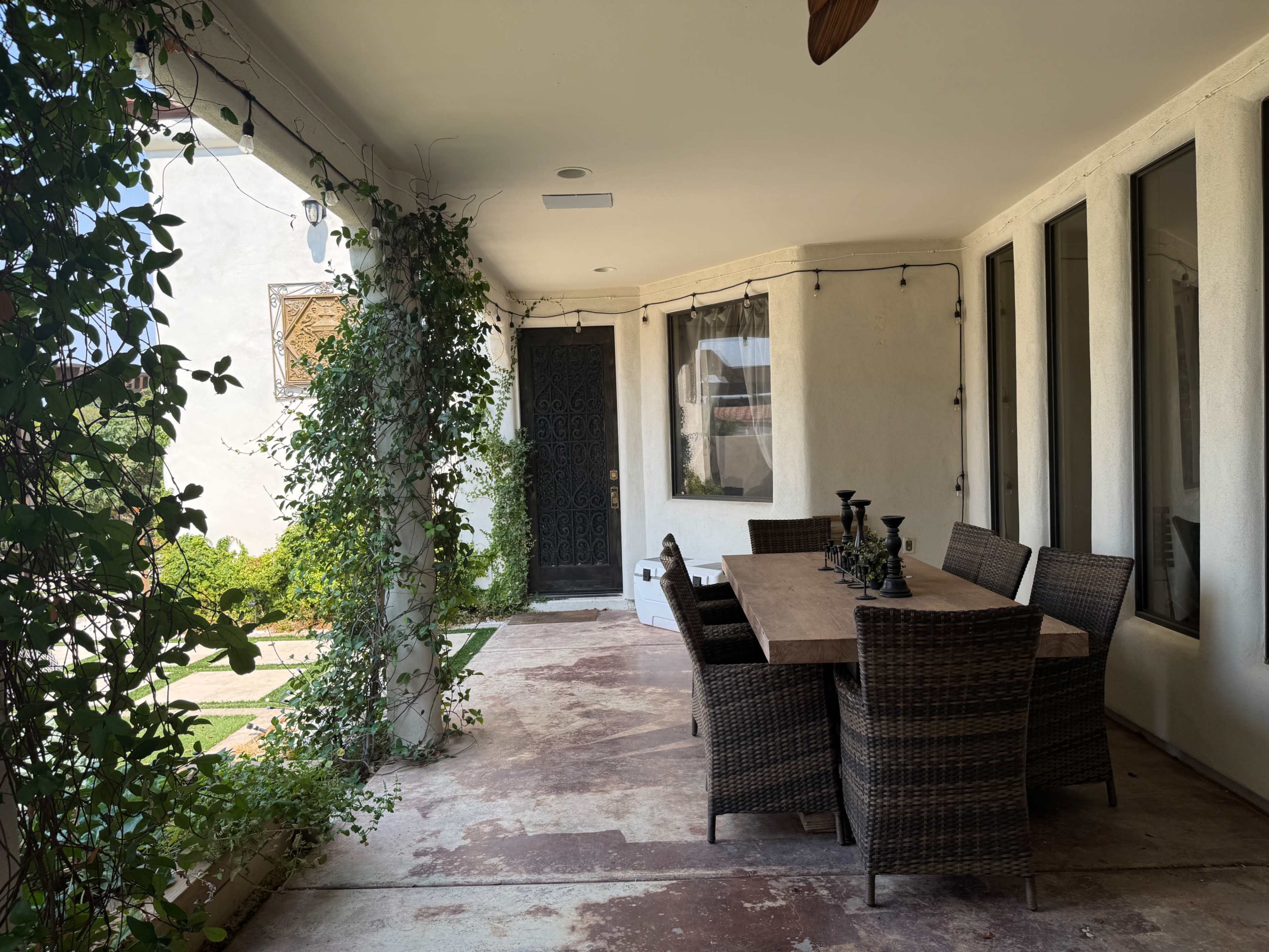 The image shows a covered outdoor patio with a large wooden dining table surrounded by six wicker chairs, and greenery along the walls.
