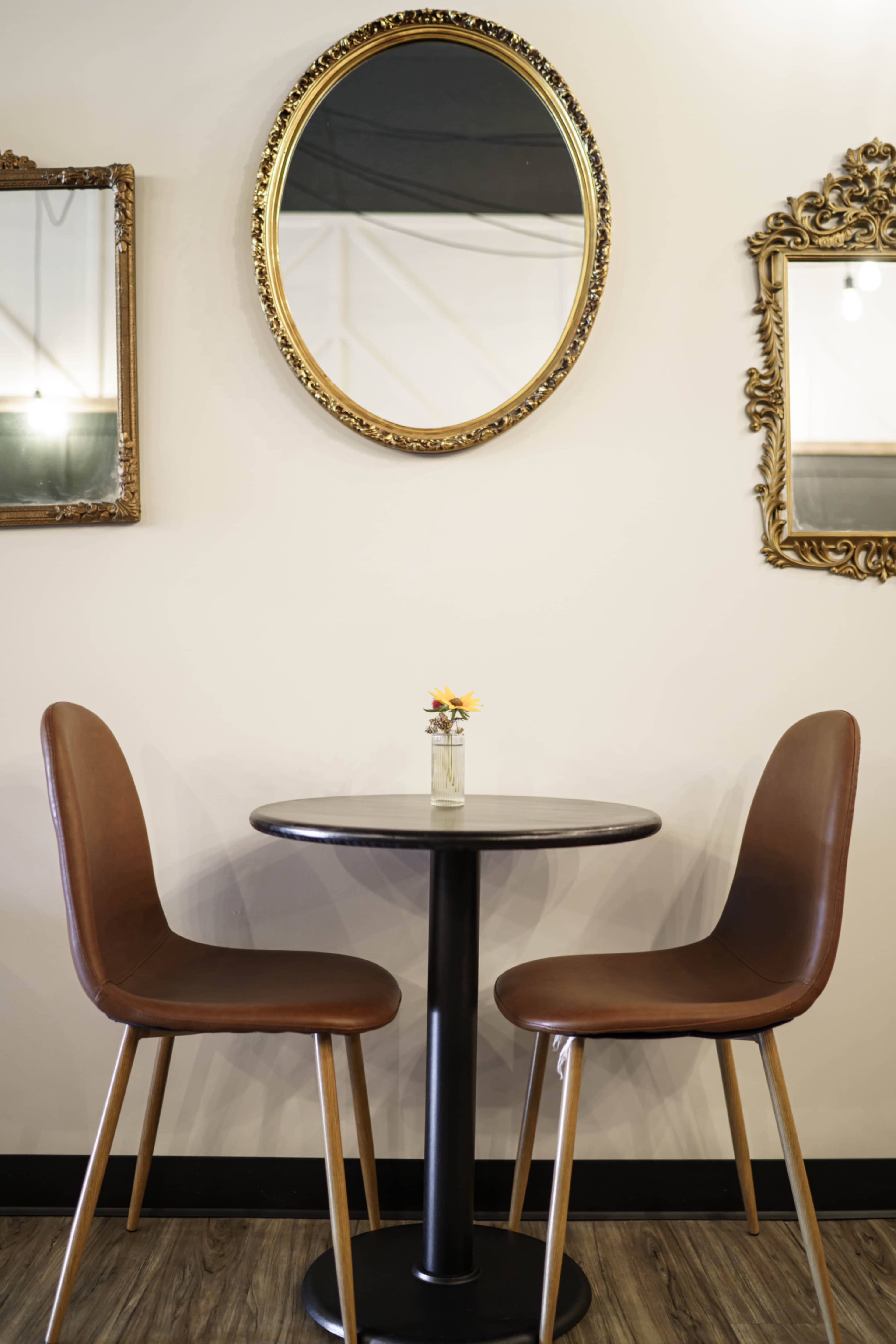 Two brown chairs are situated around a small round table, with a flower vase on top, against a wall adorned with three decorative mirrors.
