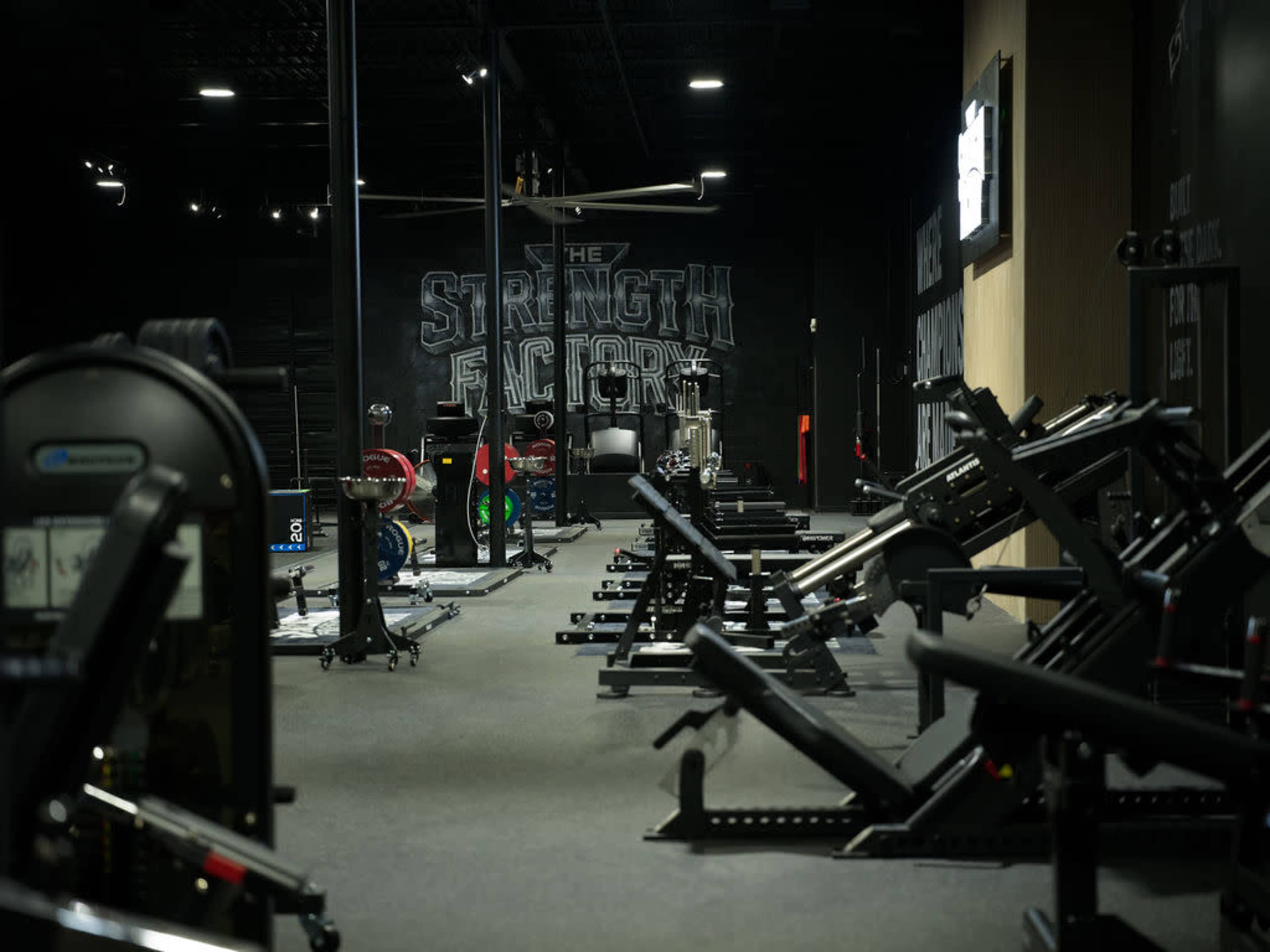 The image shows a gym interior with various weightlifting equipment and exercise machines arranged on a dark floor.