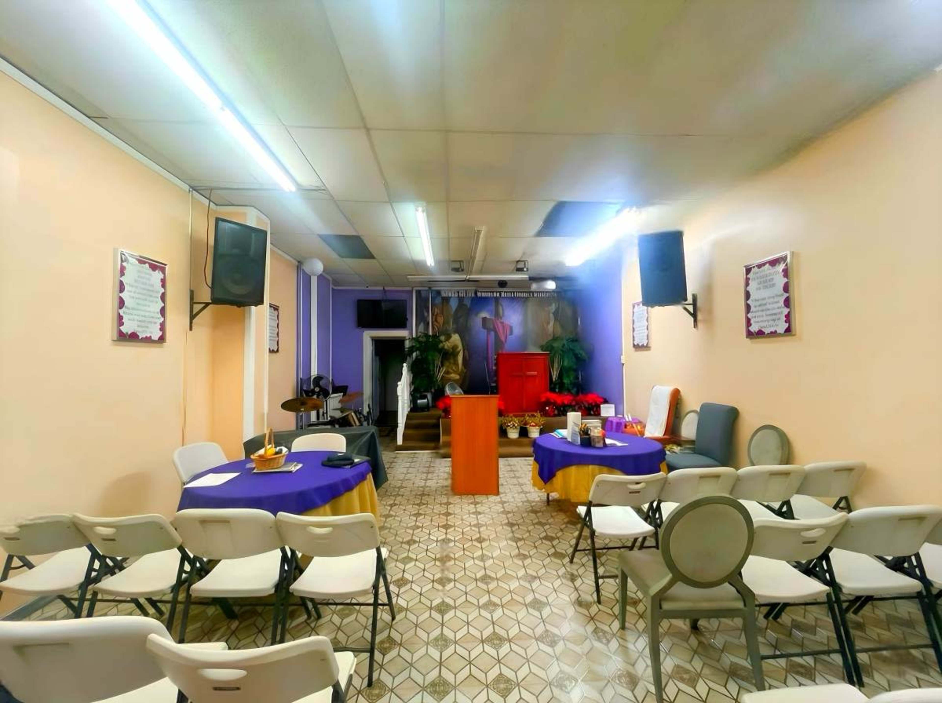 The image shows a small indoor space arranged with chairs and tables, featuring a stage or podium at one end and decorations on the walls.