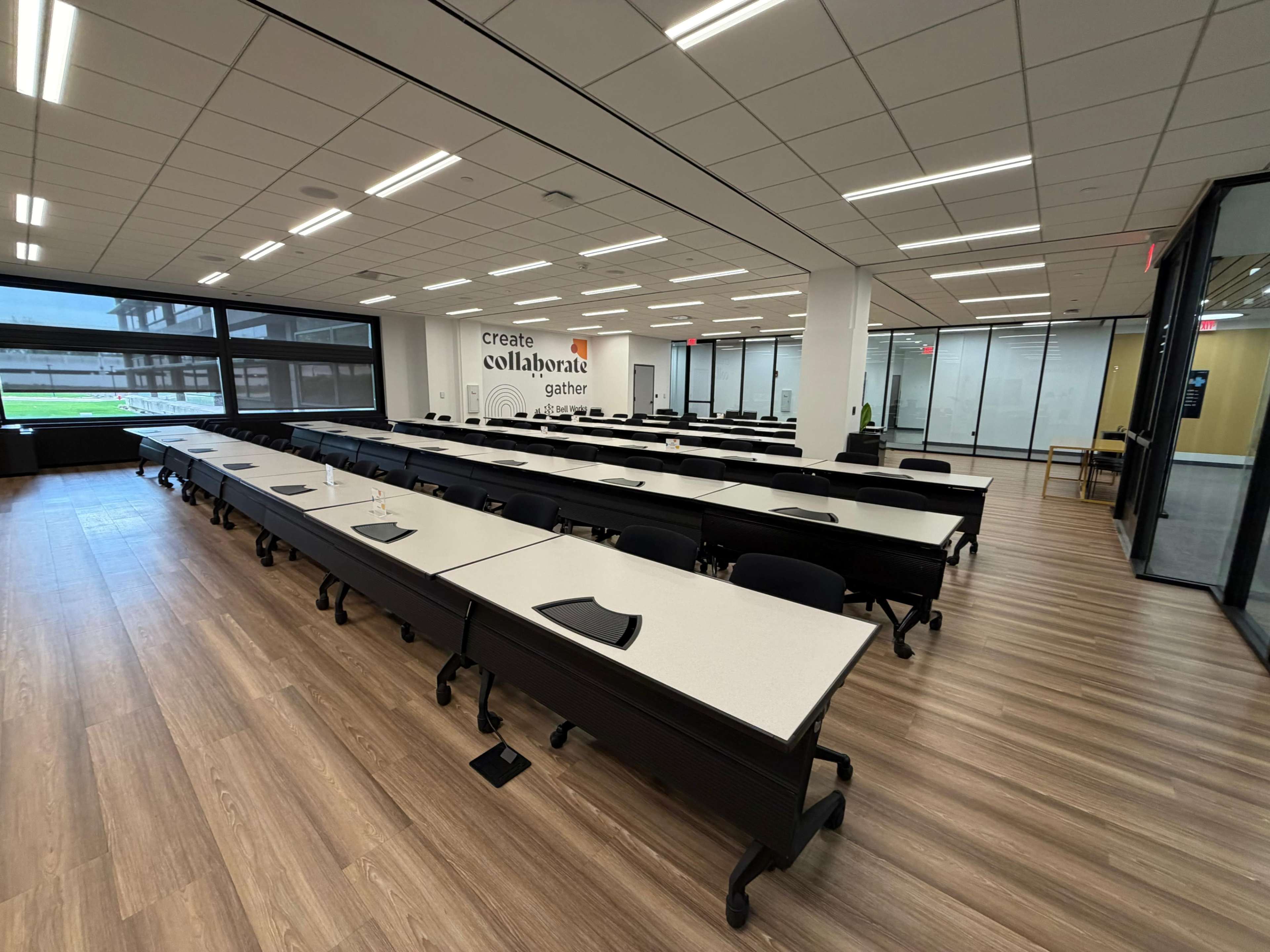 The image shows a spacious, modern classroom with rows of tables and black chairs, equipped with large windows and a whiteboard on one wall.