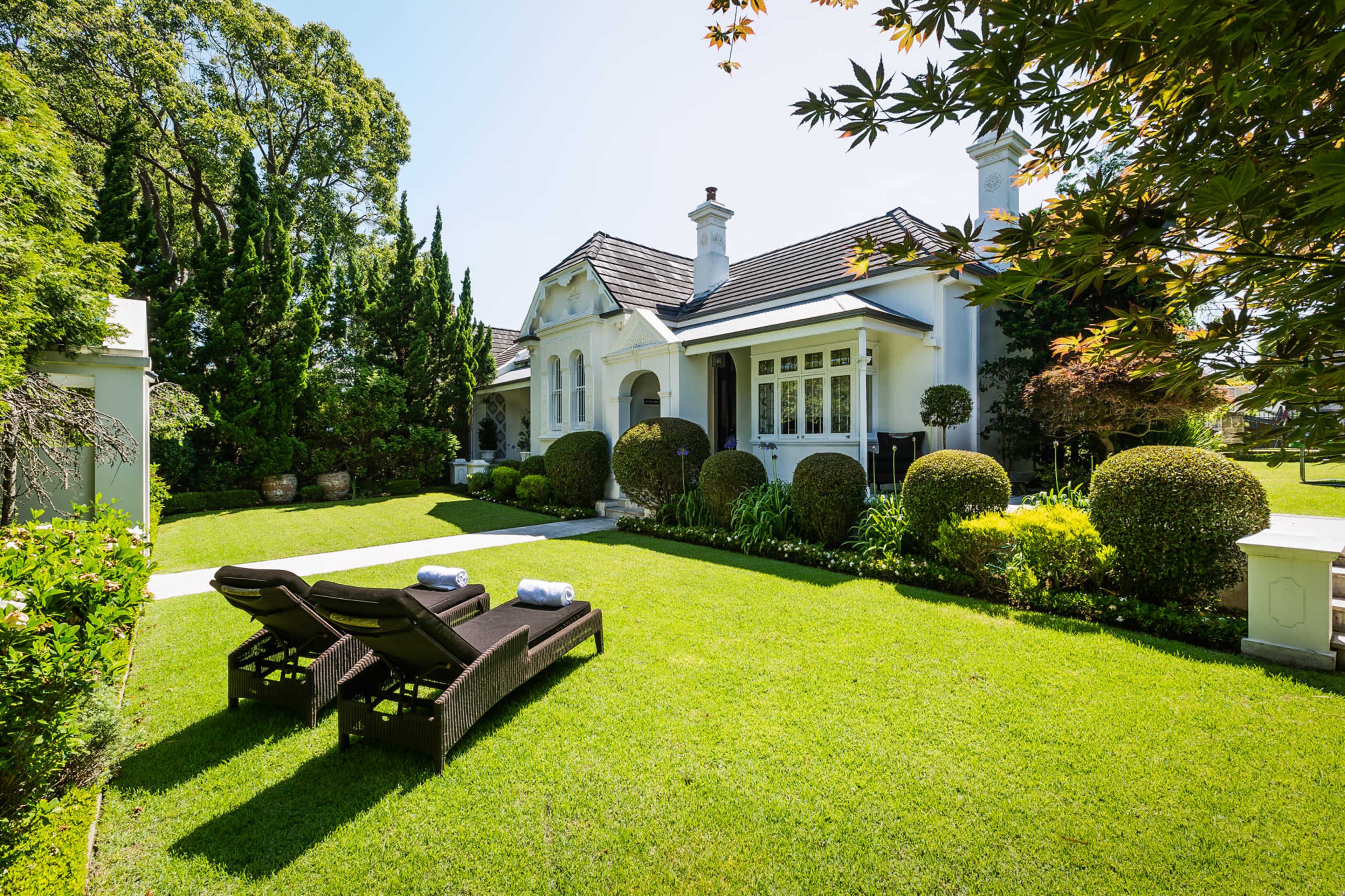 A well-manicured lawn features two lounge chairs in front of a white house surrounded by neatly trimmed hedges and various trees.