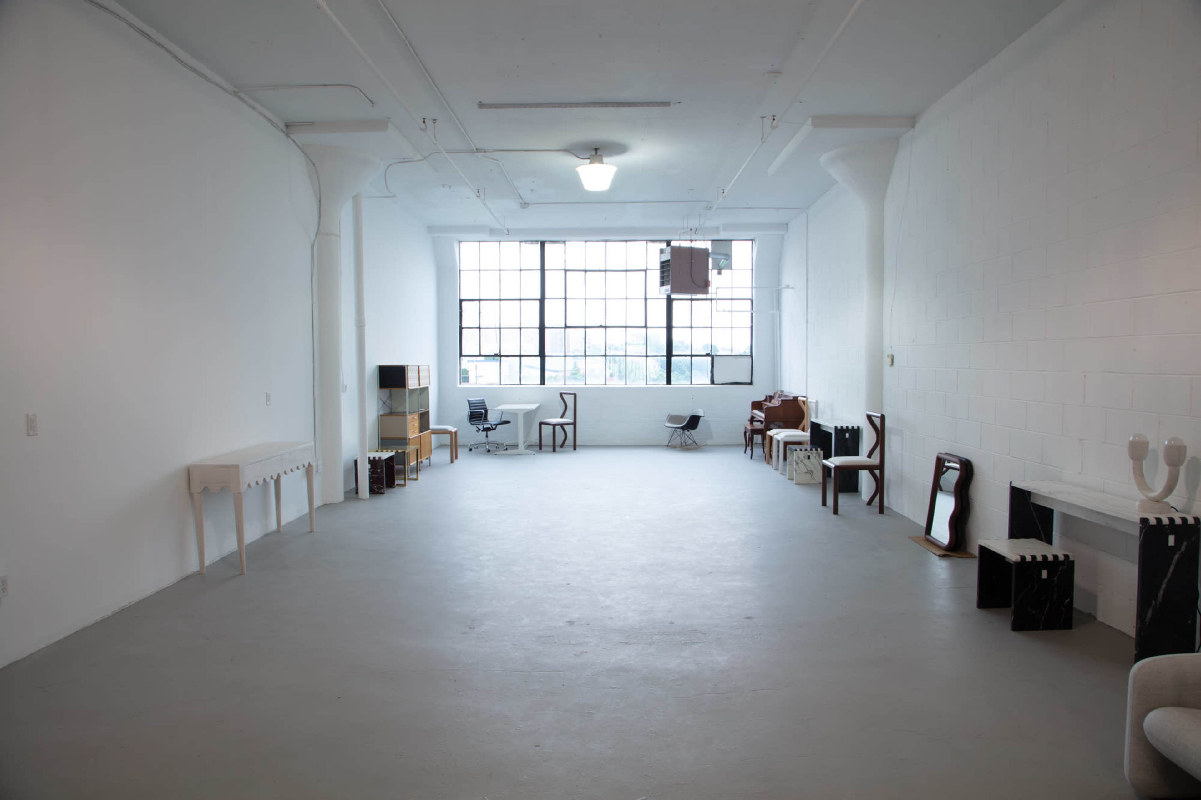 A spacious, empty room with concrete flooring and large windows, furnished with various pieces of furniture along the walls.