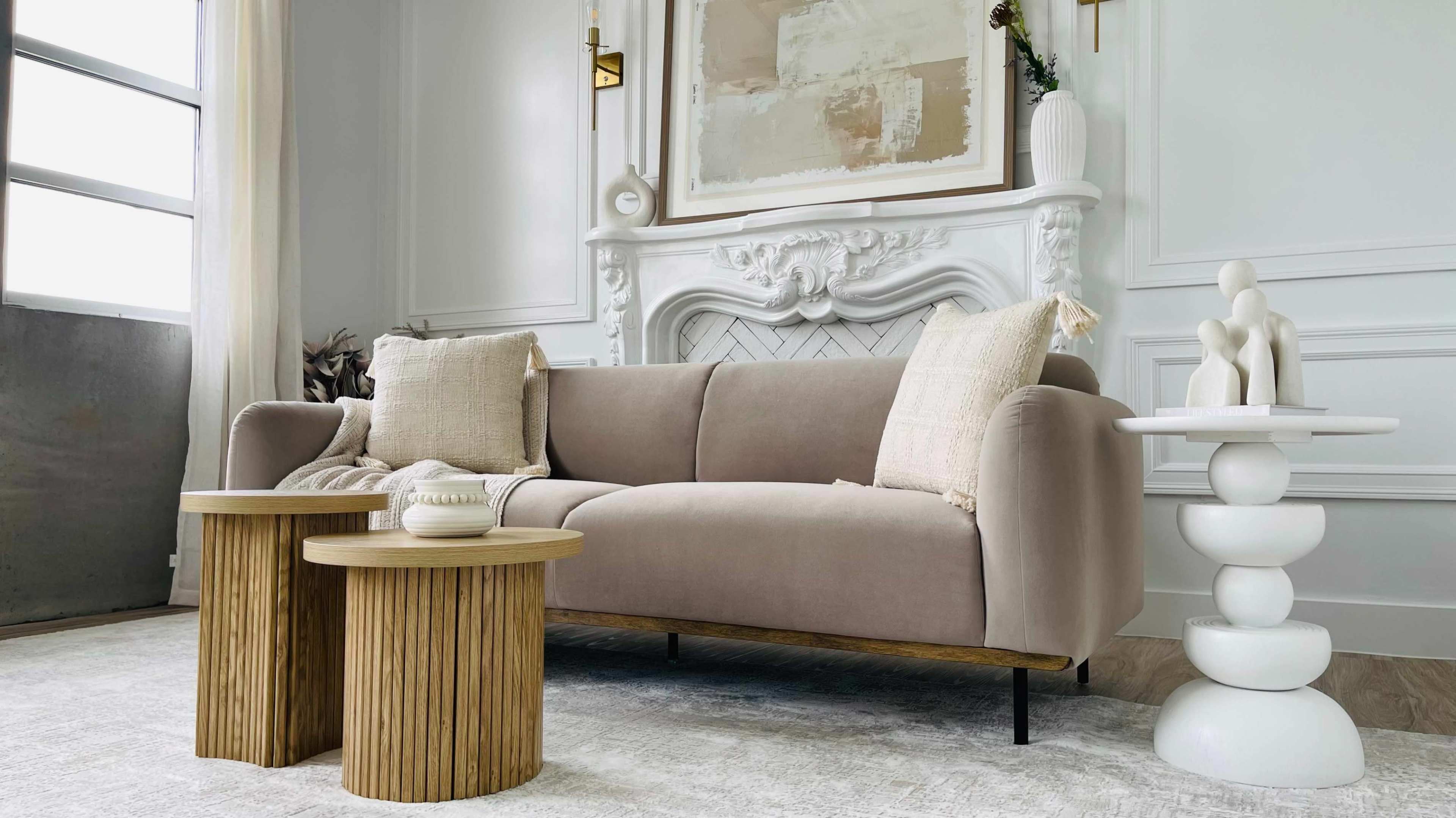 The image shows a modern living room featuring a beige sofa, two round wooden side tables, and a decorative white sculpture on a pedestal next to the sofa.