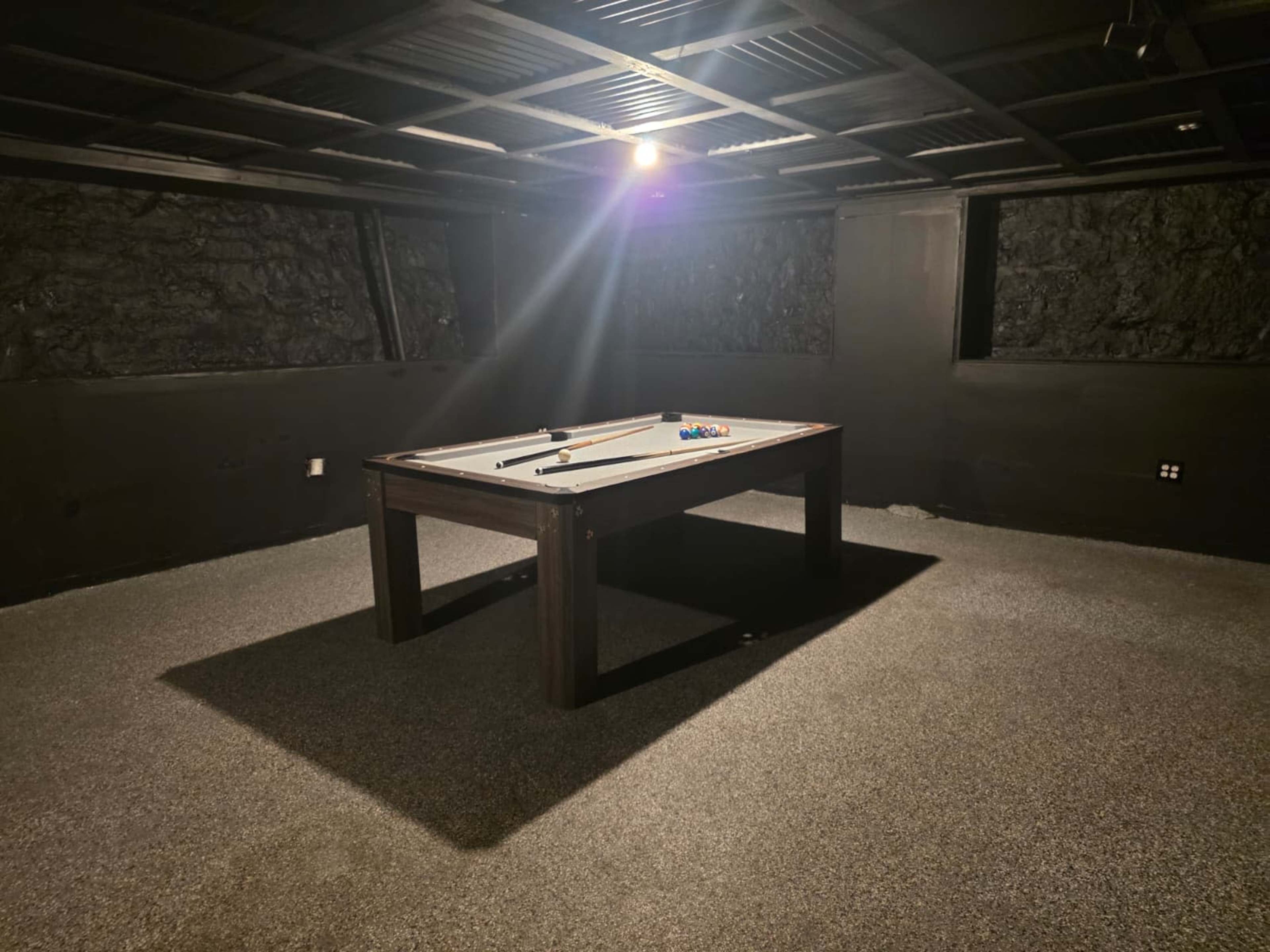 A pool table stands in the center of a dimly lit room with dark walls and carpet.
