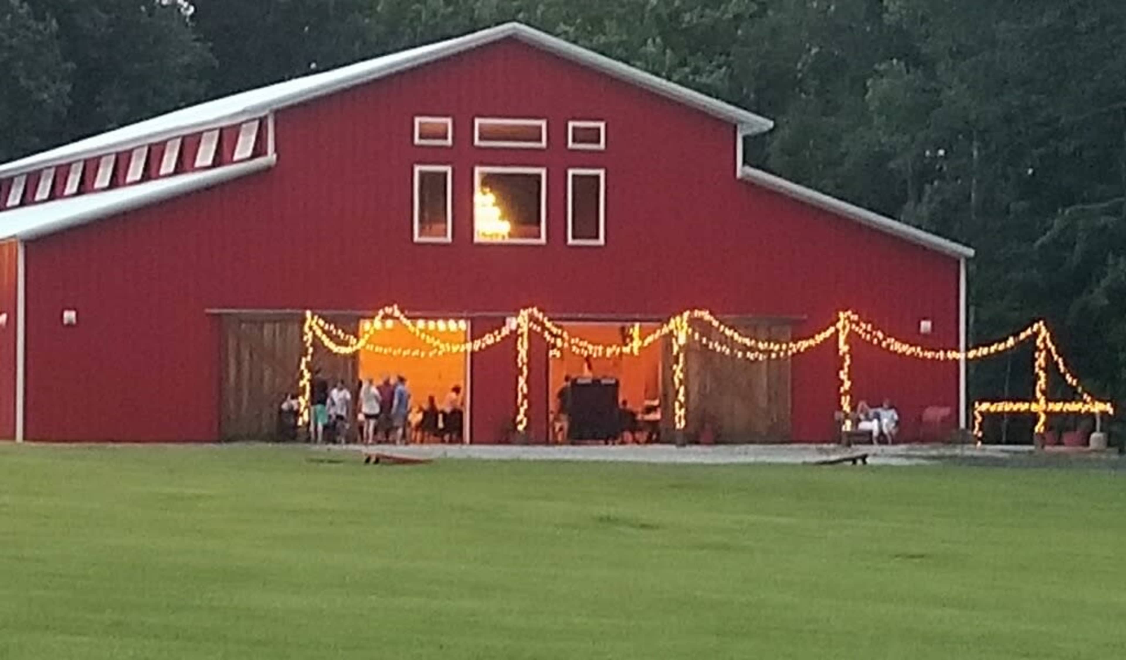 A red barn with bright string lights is partially open, revealing a gathering of people inside as the sun sets.