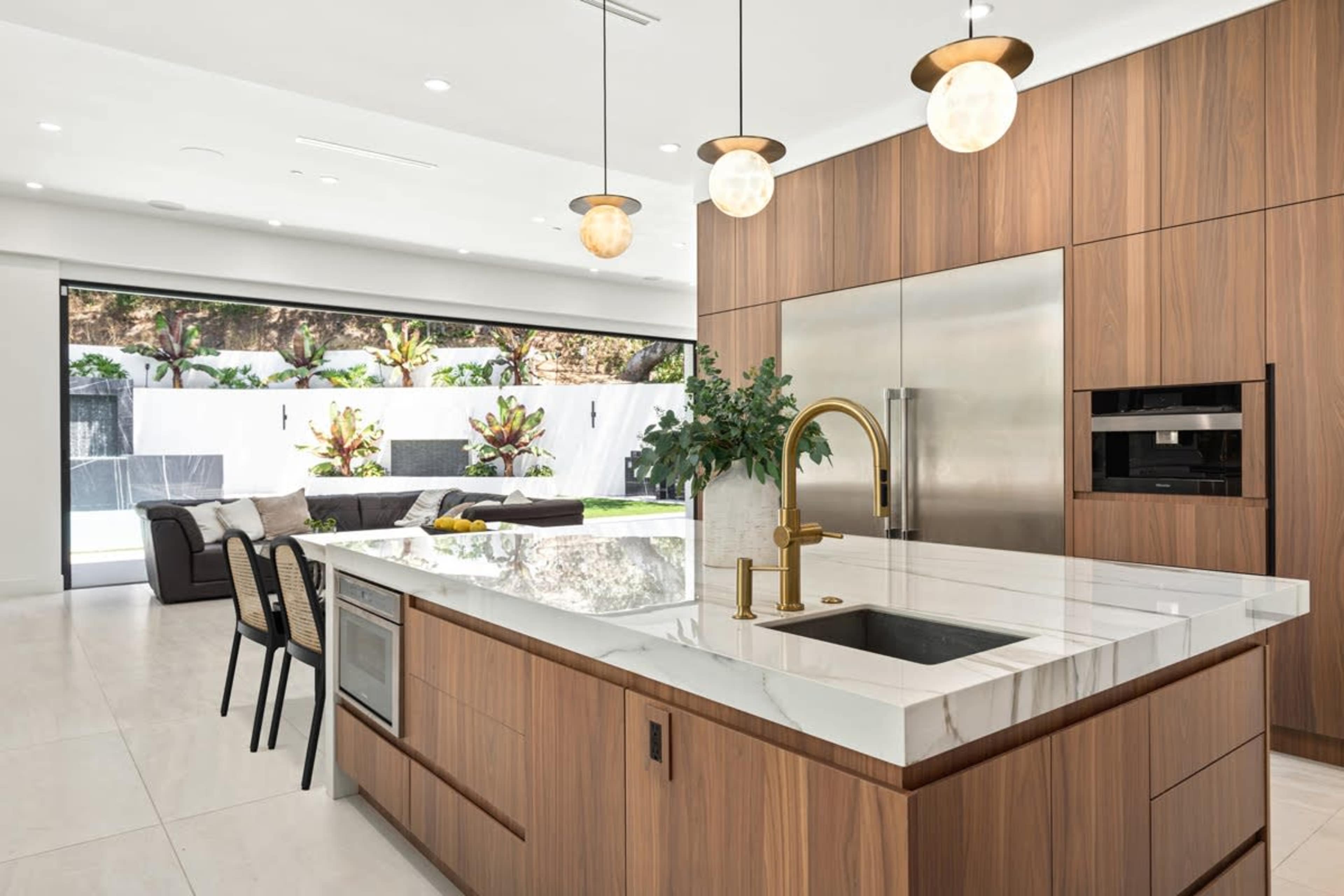 A modern kitchen with wooden cabinetry, a large marble island, and sleek appliances, connected to a patio visible through large glass doors.