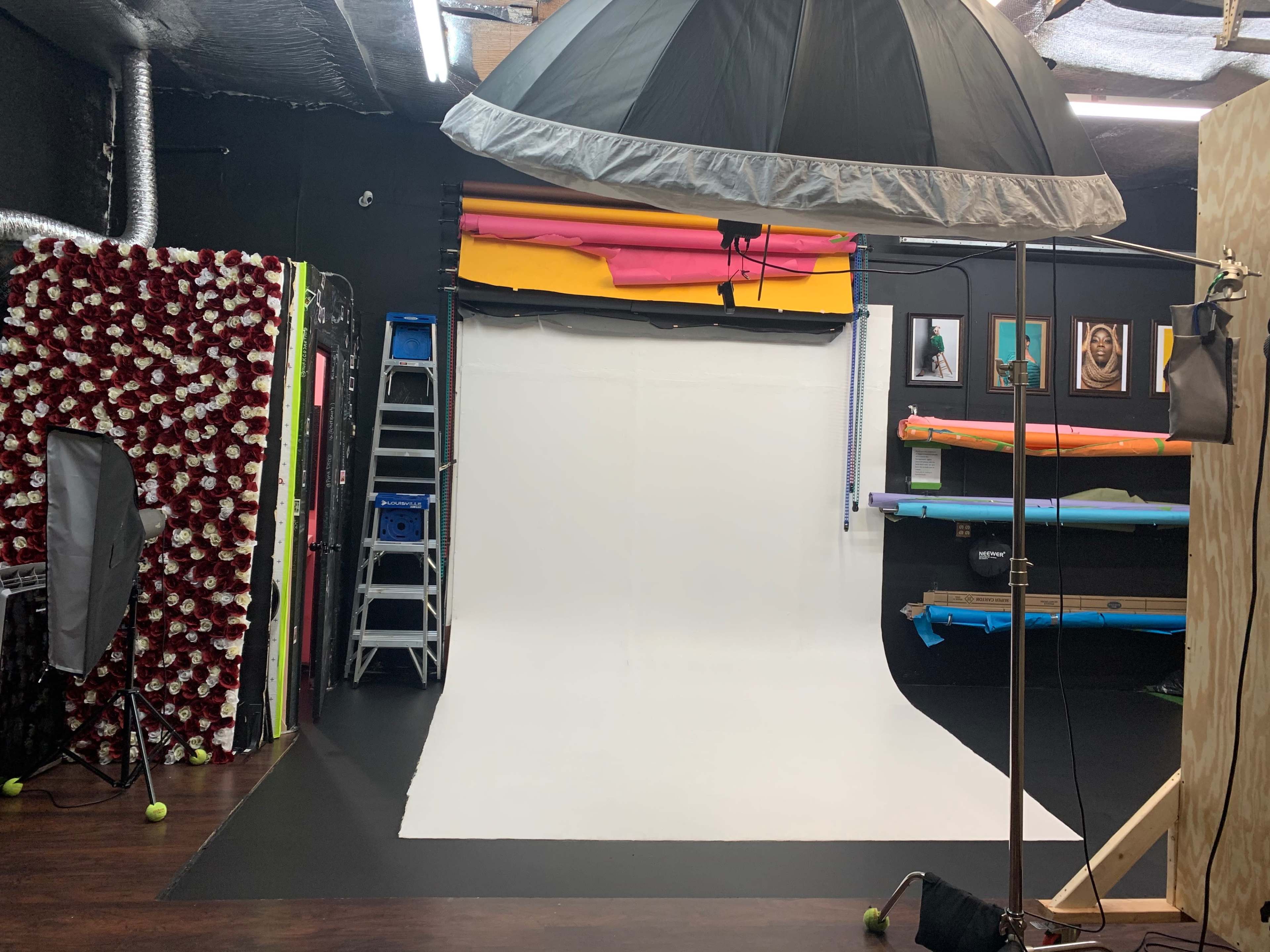 A photography studio with a white backdrop, various colored backdrops stacked on shelves, and lighting equipment set up.