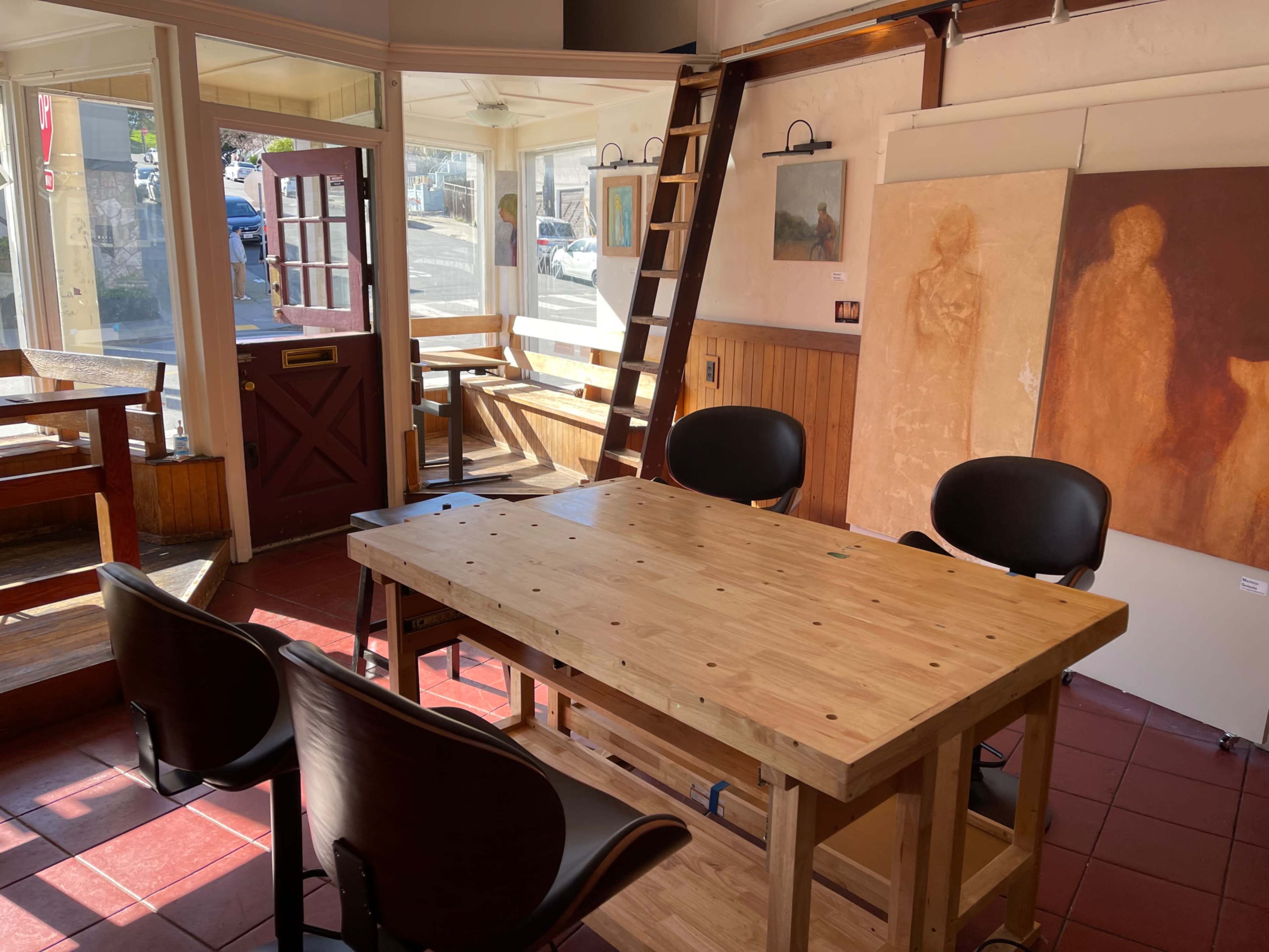 The image shows a sunlit art studio with a wooden table and chairs, featuring a ladder and paintings on the walls, along with a view of the street through a door.
