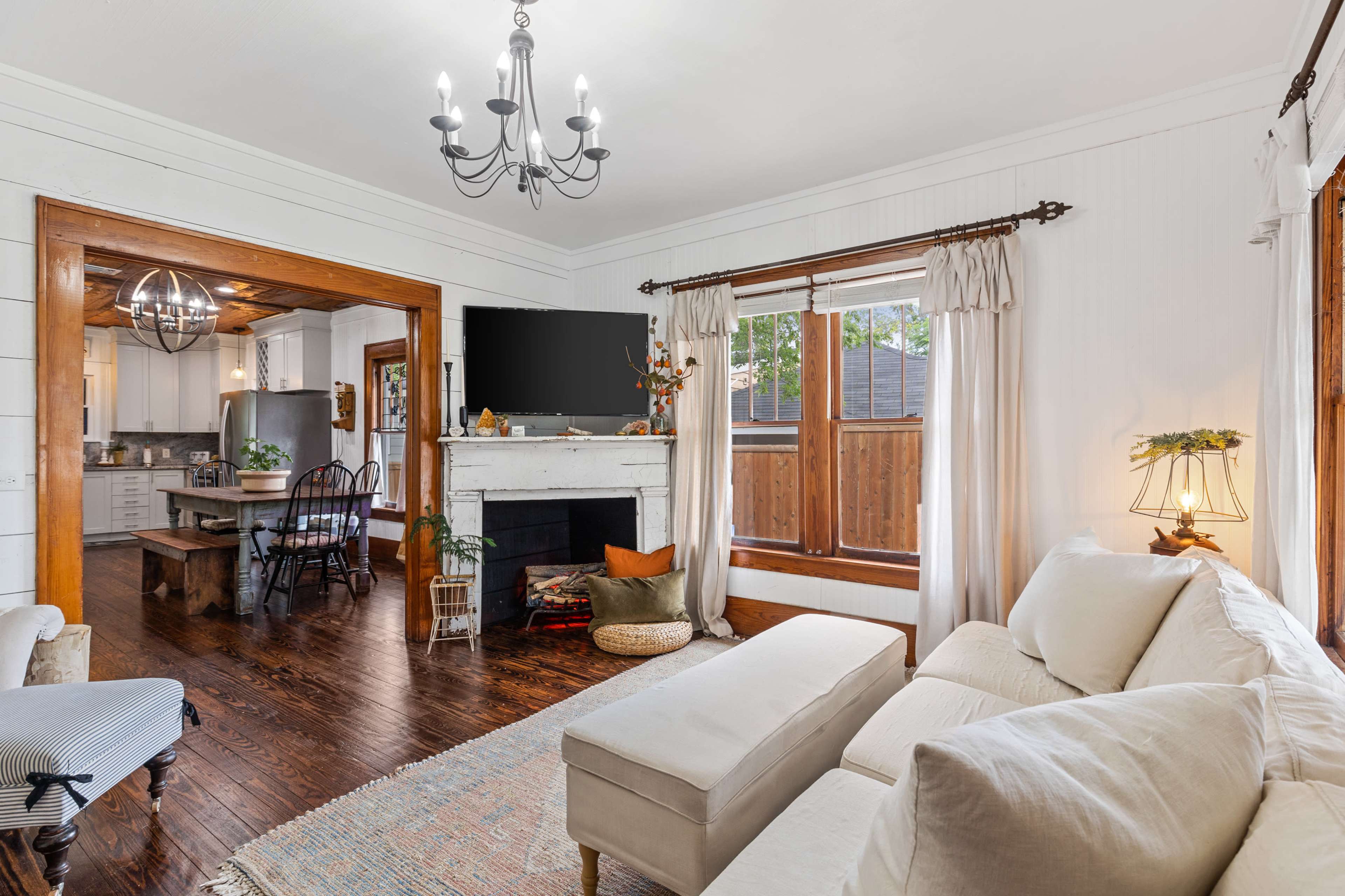 A cozy living room with a sectional sofa, a coffee table, and large windows letting in natural light, alongside a fireplace and a dining area visible in the background.