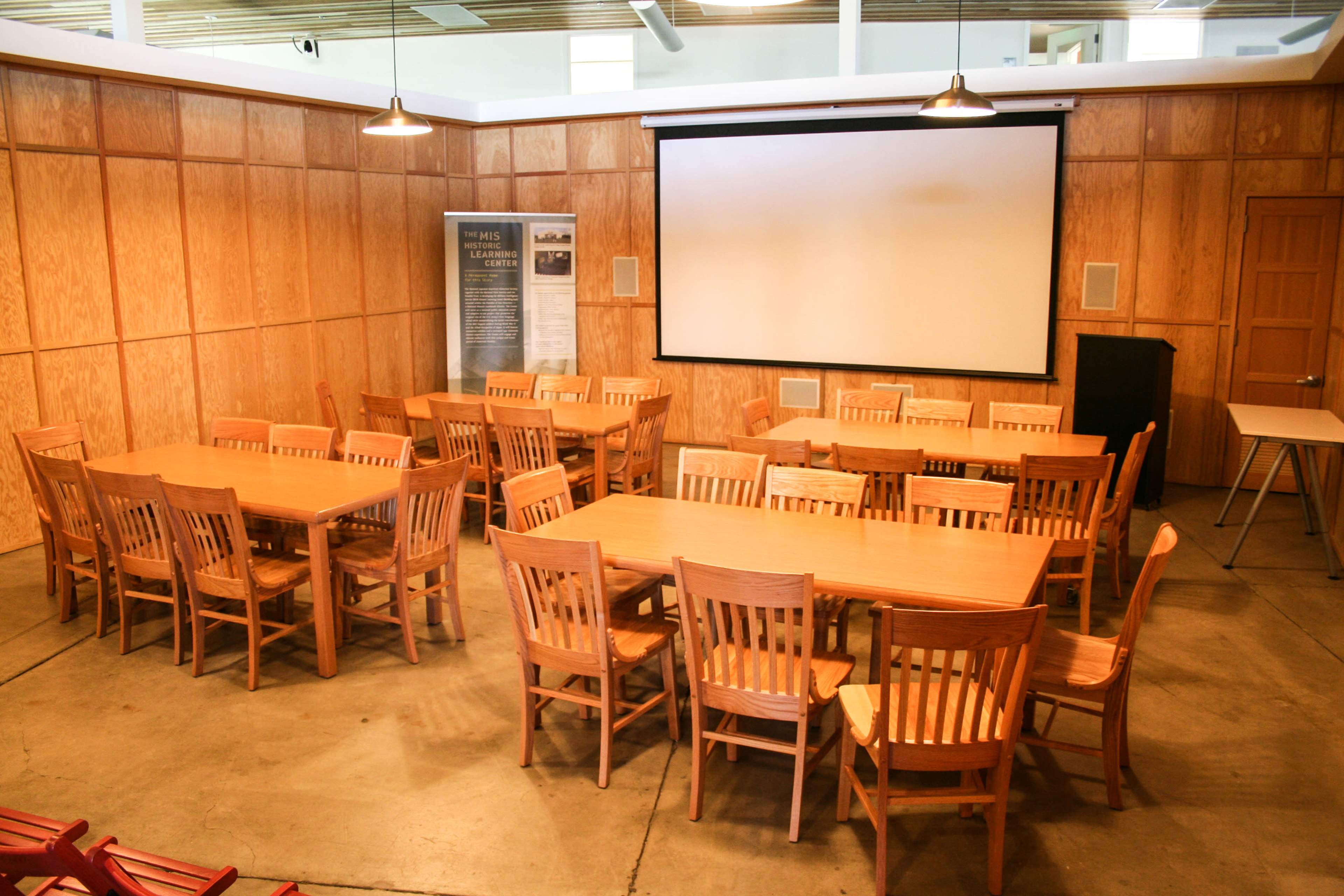 The image shows a spacious, wooden-furnished room arranged with multiple rectangular tables and chairs, centered around a projector screen.