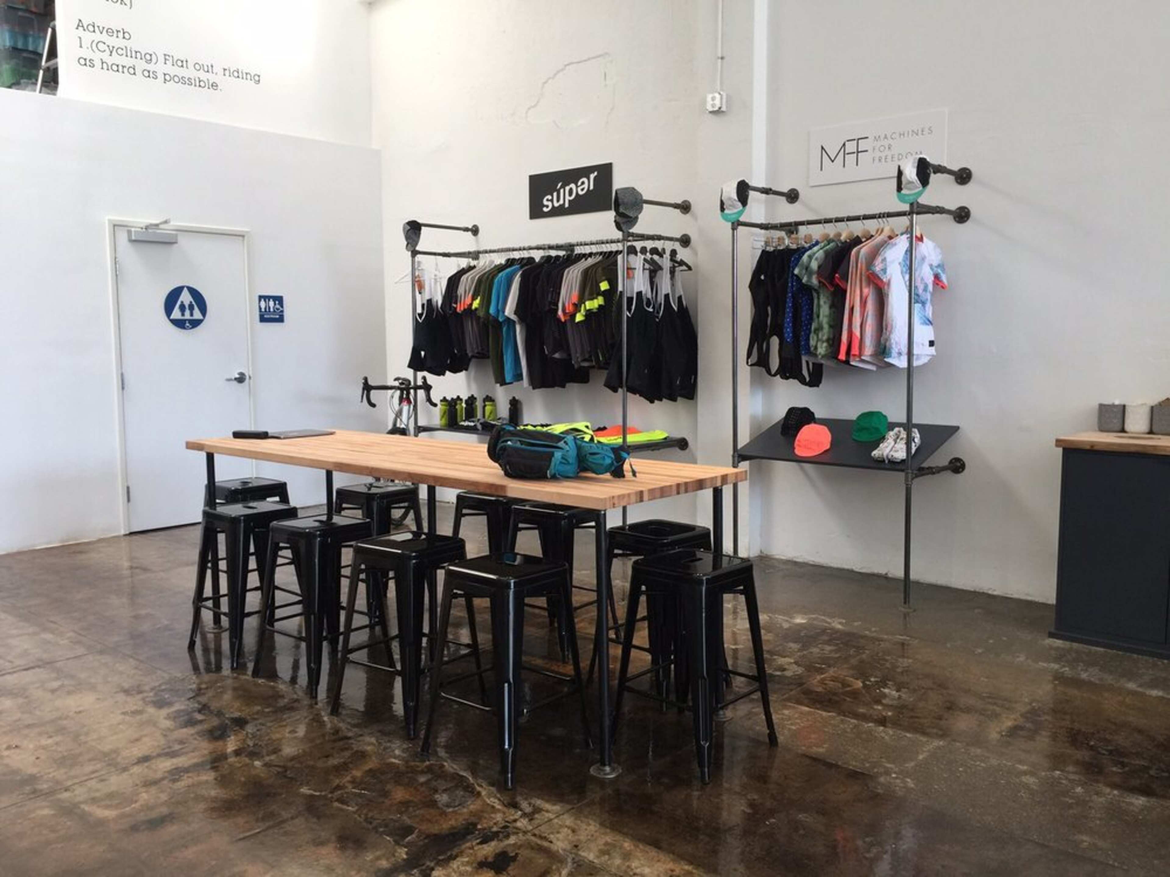 A clothing display area with racks of athletic wear and a wooden table surrounded by metal stools in a minimalist room.