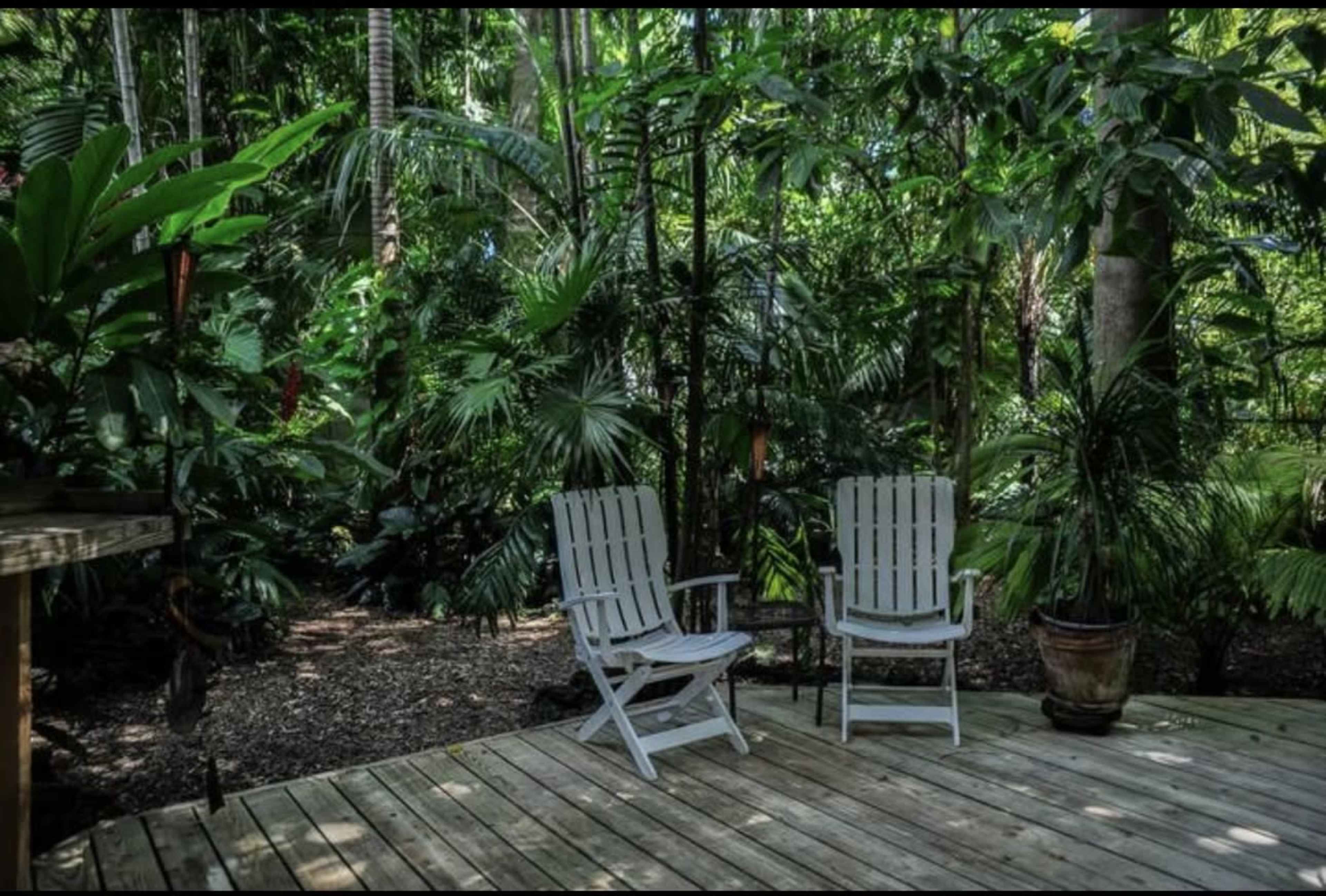 A wooden deck features two white Adirondack chairs surrounded by lush tropical greenery.