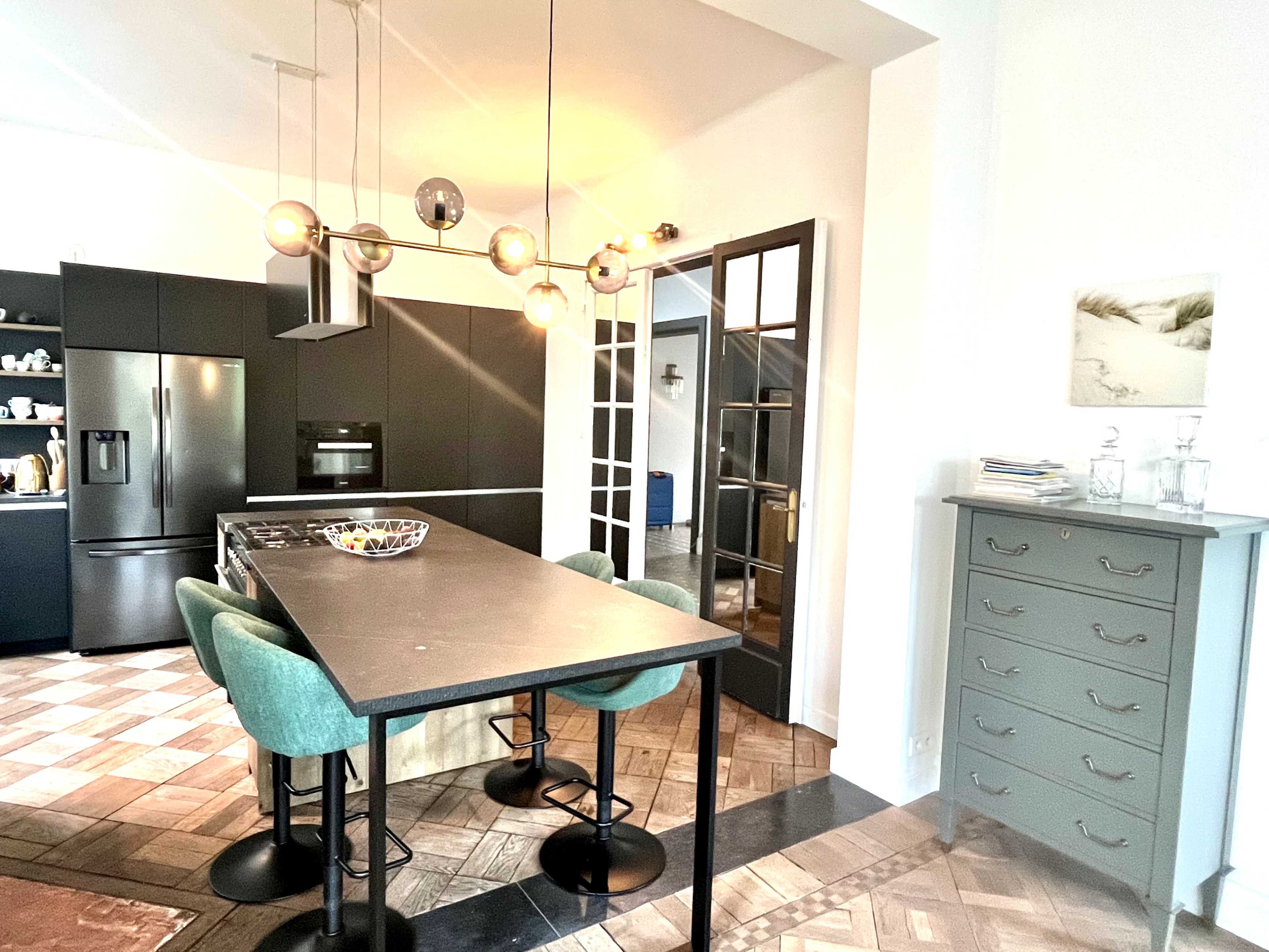 A modern kitchen features a black island with green chairs, pendant lights overhead, and sleek cabinetry in the background.
