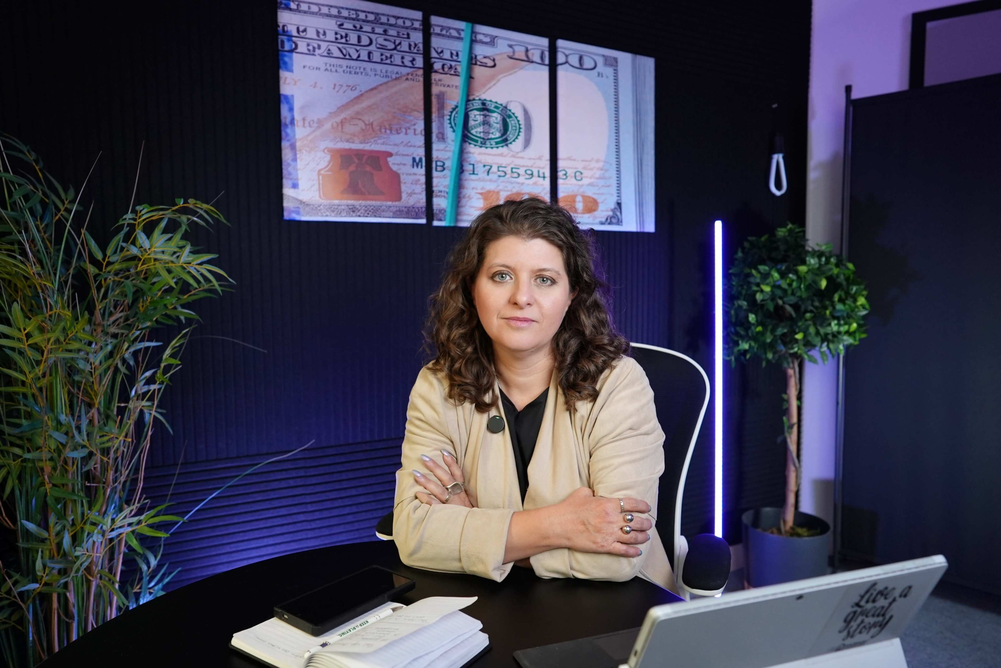 A woman sits at a desk in a modern office space, with a large image of a hundred-dollar bill displayed on the wall behind her.