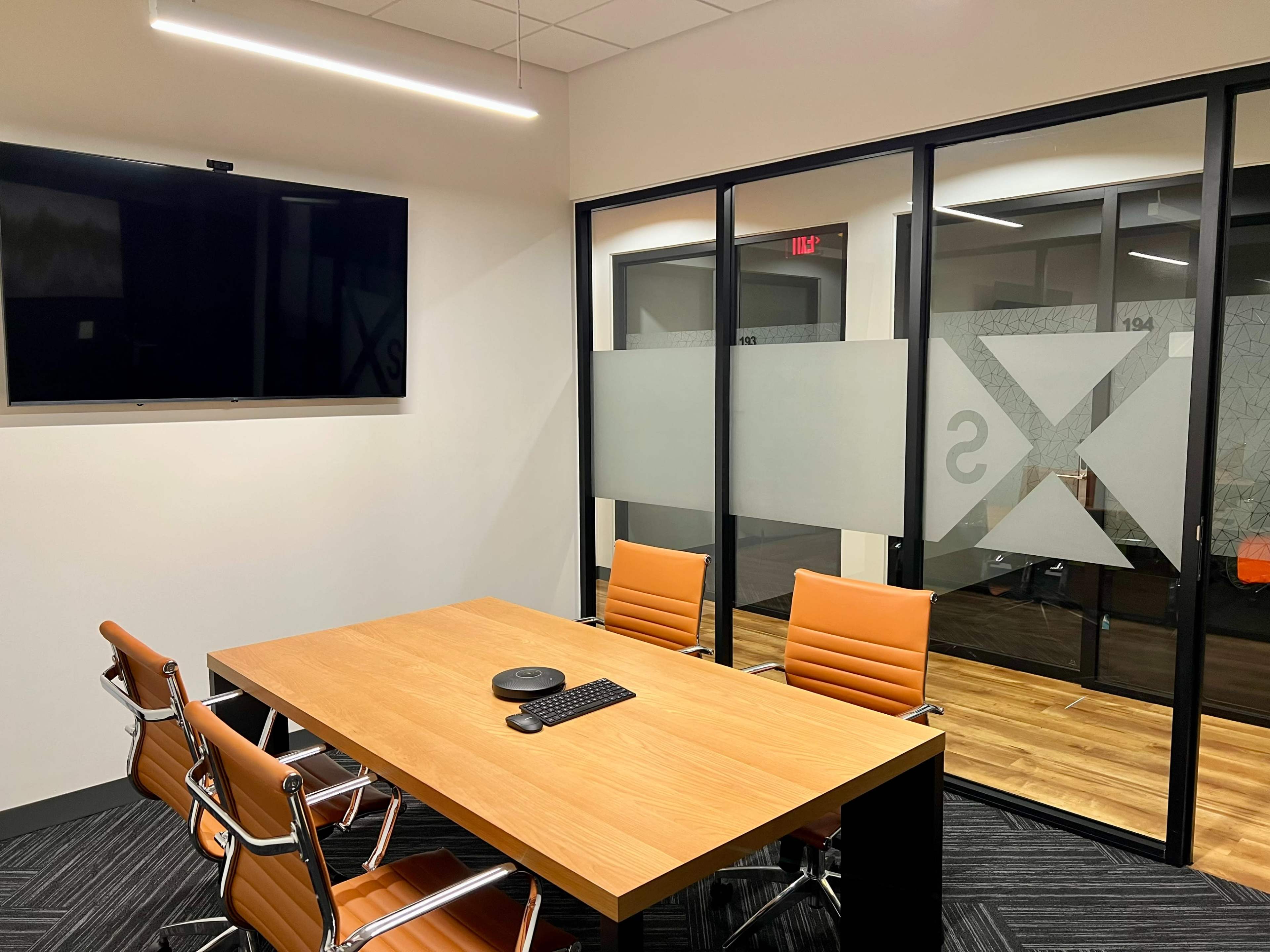 The image shows a modern conference room with a wooden table, four orange chairs, and a wall-mounted TV.