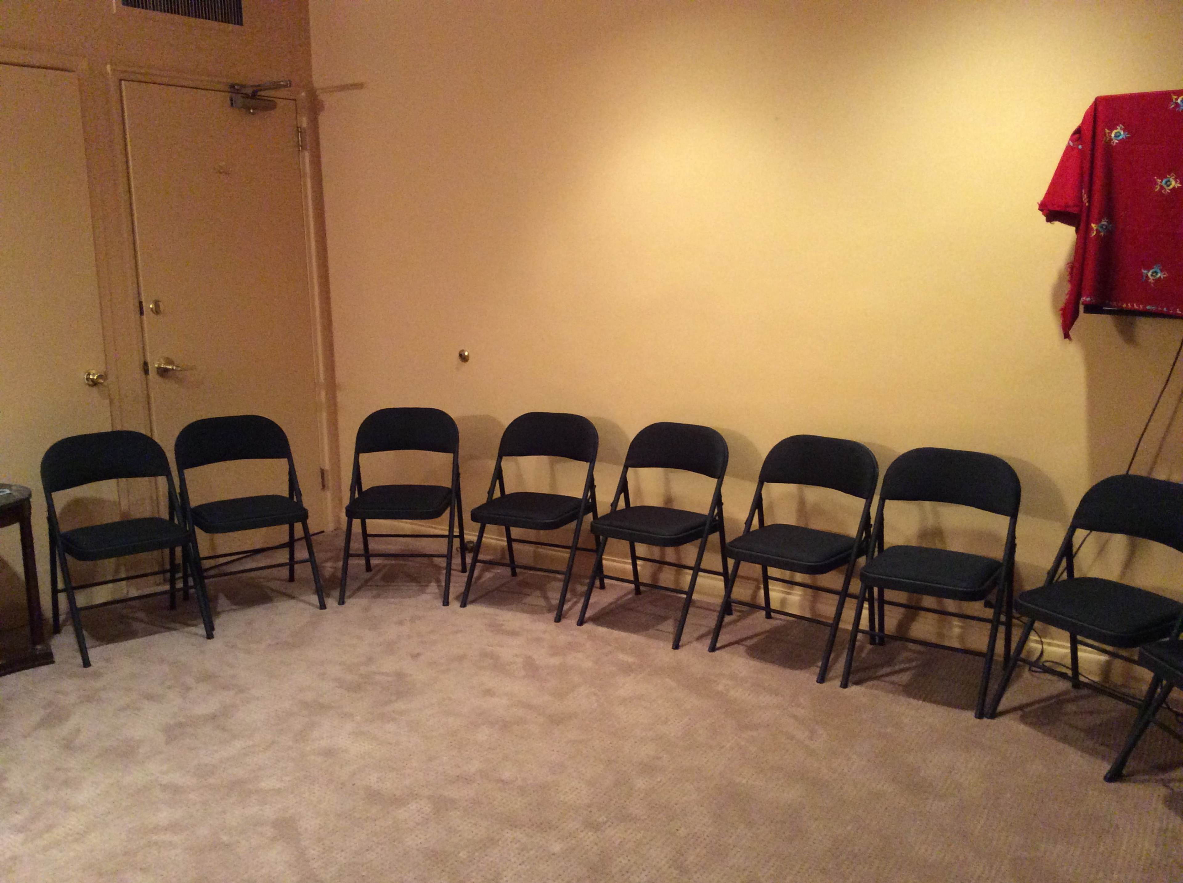 A row of eight black folding chairs is arranged against a beige wall in a carpeted room.