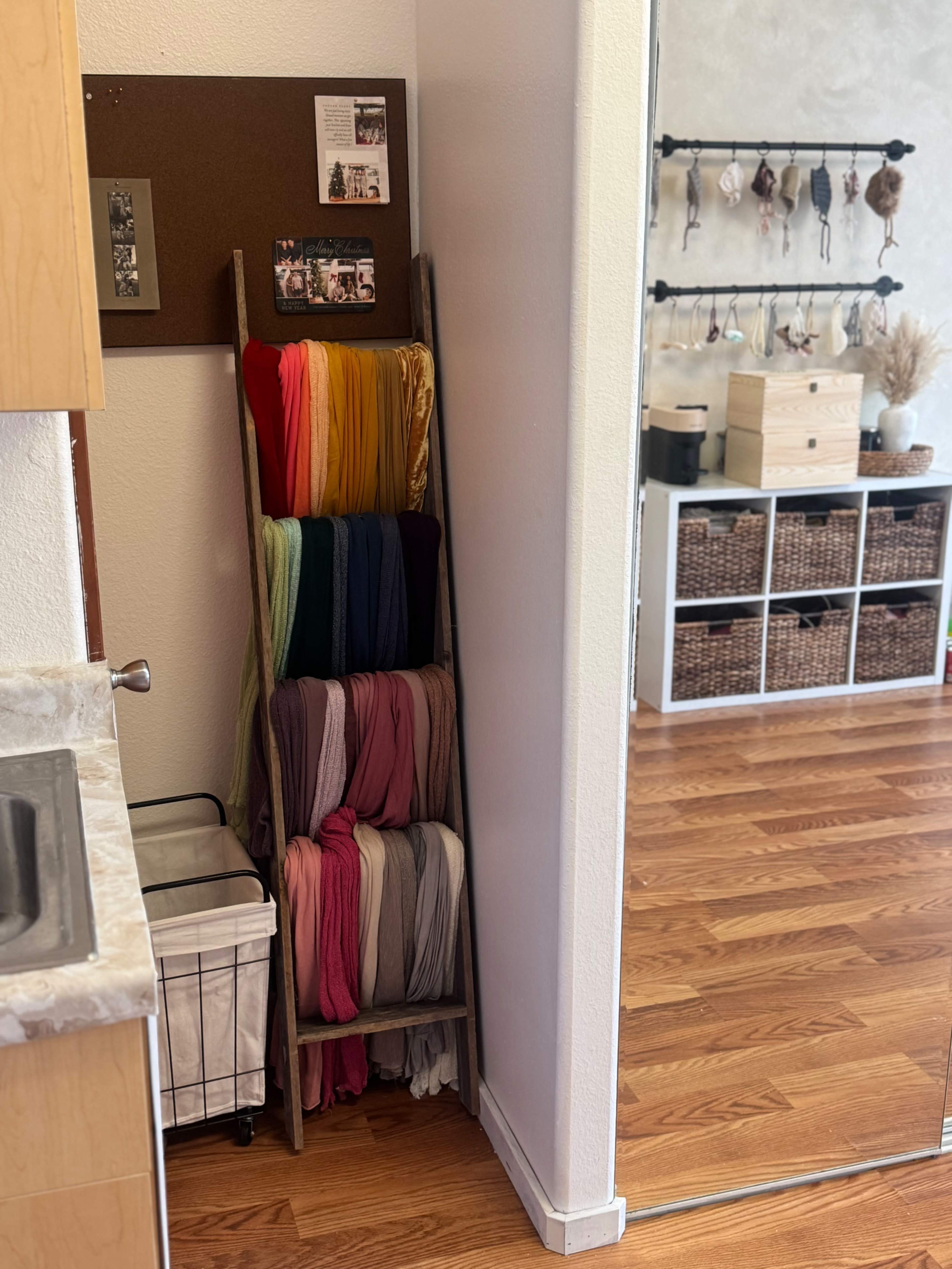 The image shows a corner of a room featuring a wooden rack holding colorful fabric rolls beside a sink and a view of woven storage baskets in an adjacent area.