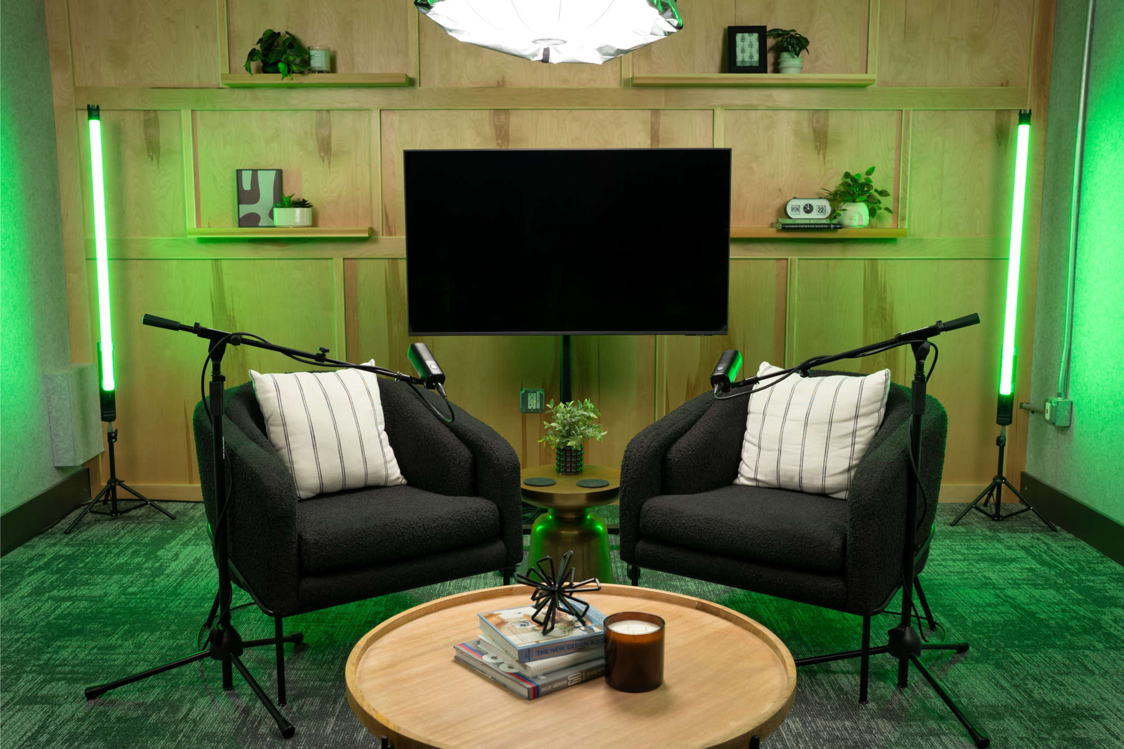 The image shows a cozy interview setup with two plush chairs, a coffee table, and a large television screen, surrounded by green accent lighting and decorative plants.
