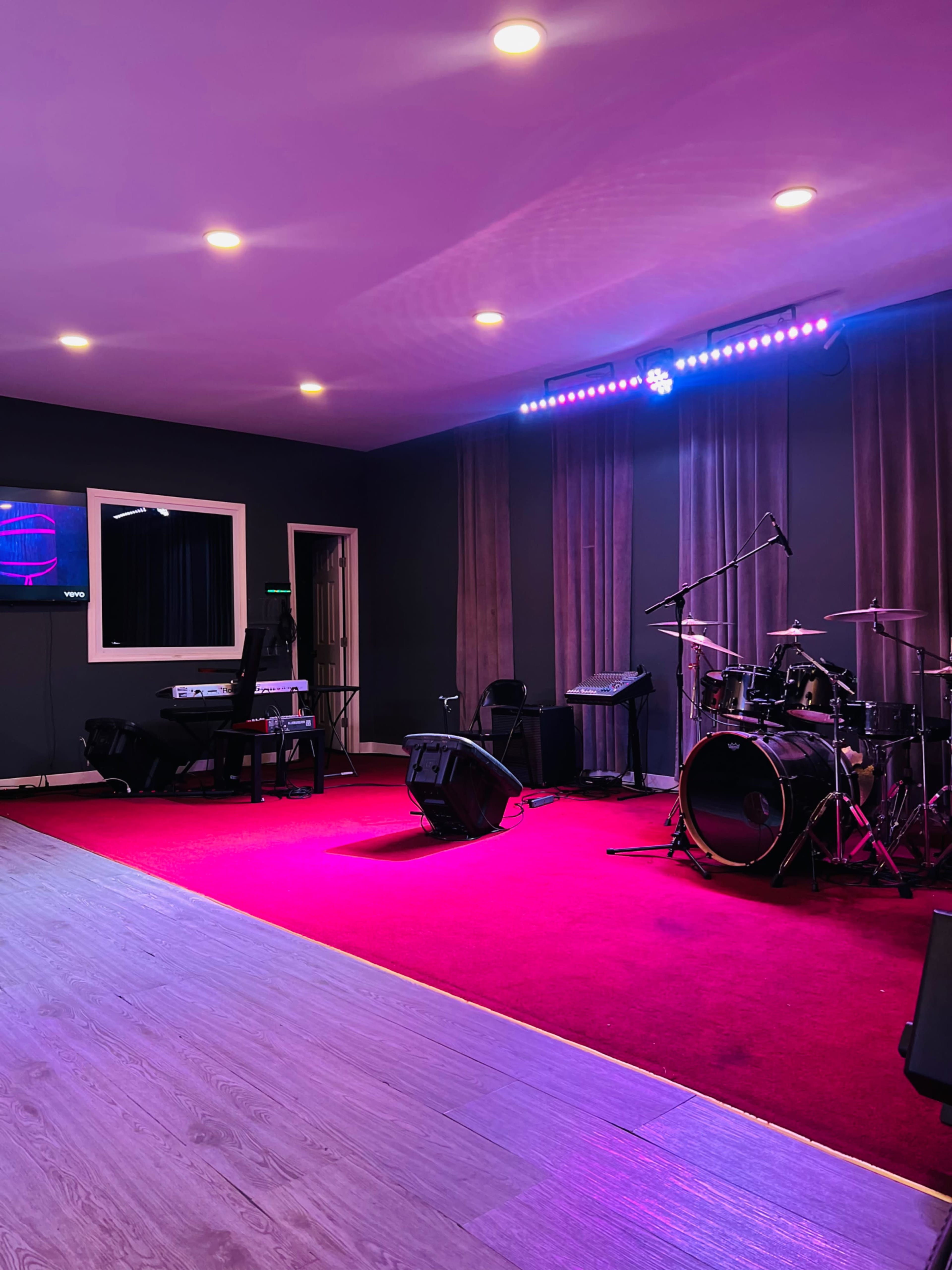 The image shows a music stage setup with a drum kit, keyboard, and amplifiers on a red carpeted floor, illuminated by purple and white stage lights.
